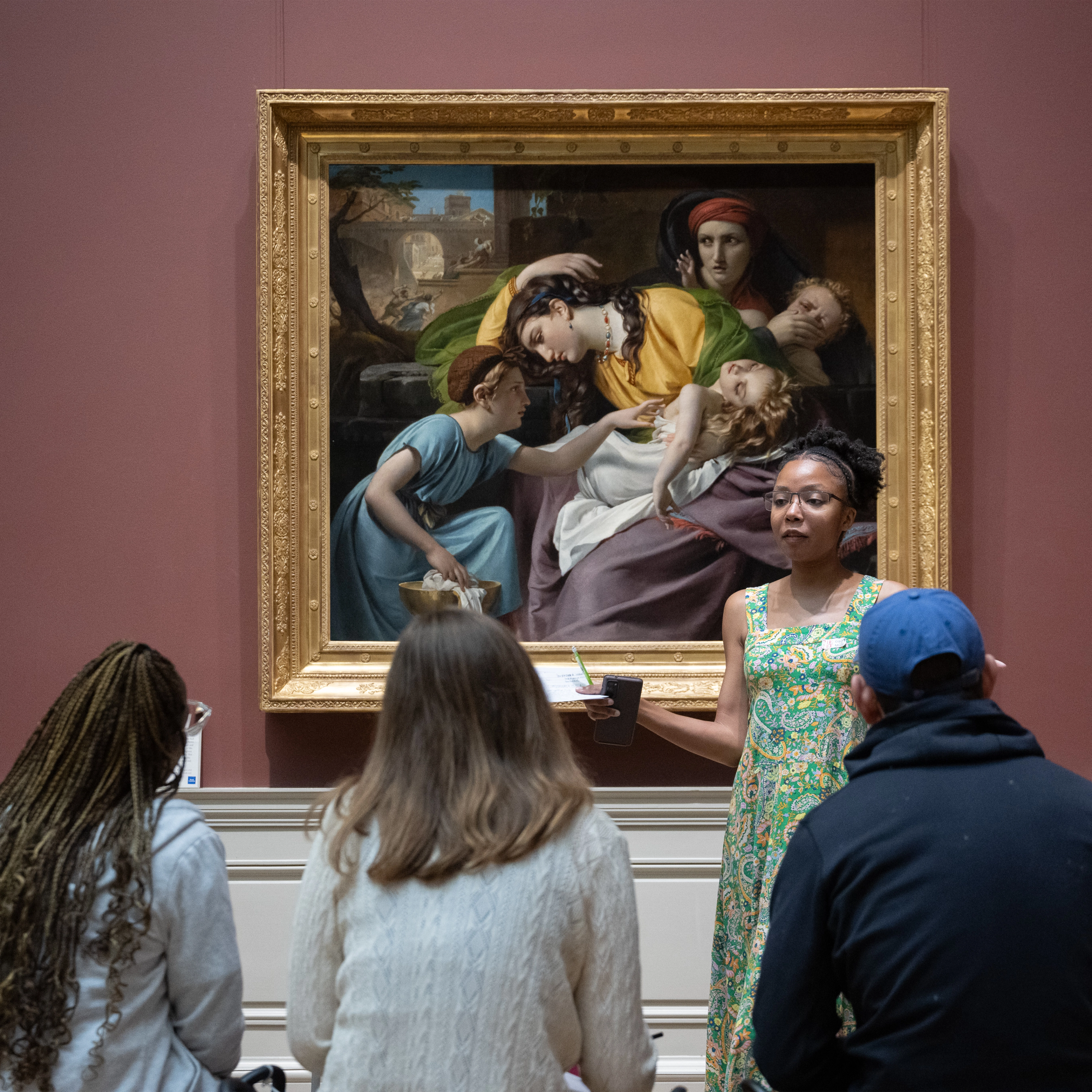 A group of people sit in front of an artwork. The woman in the center, wearing green dress, is discussing the art as the others listen.