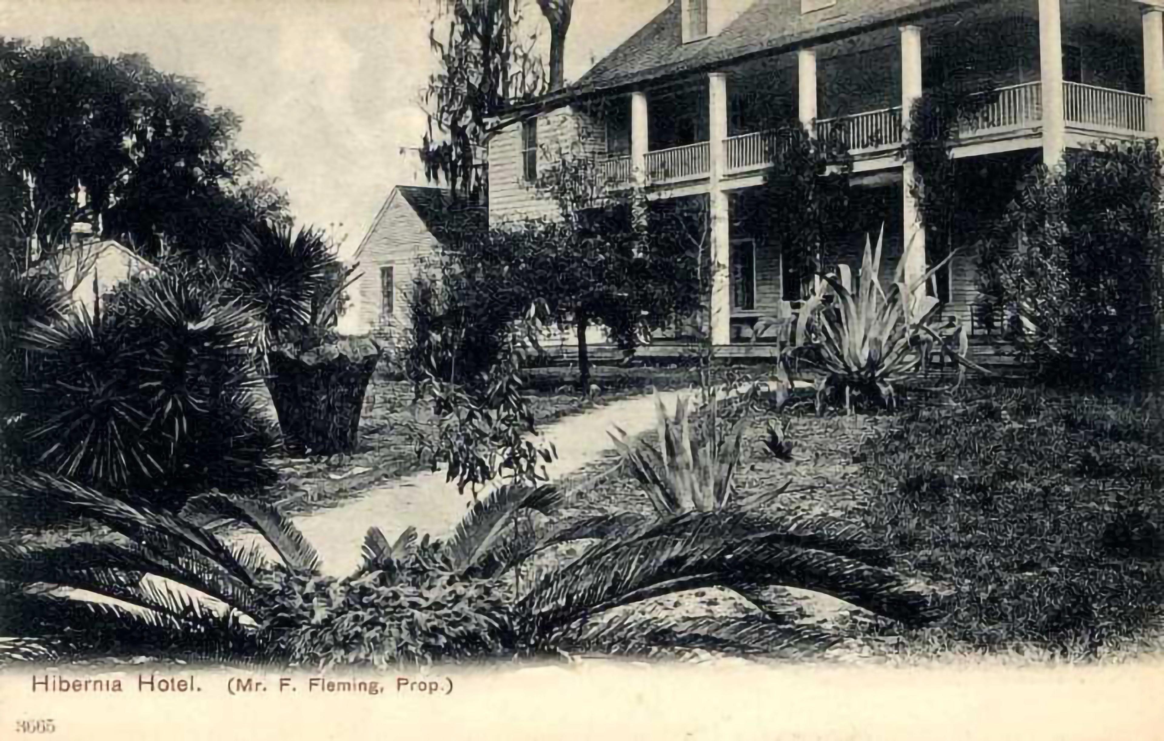 Black-and-white photograph of a large hotel with wraparound porch, surrounded by lush landscaping and a curved garden path.