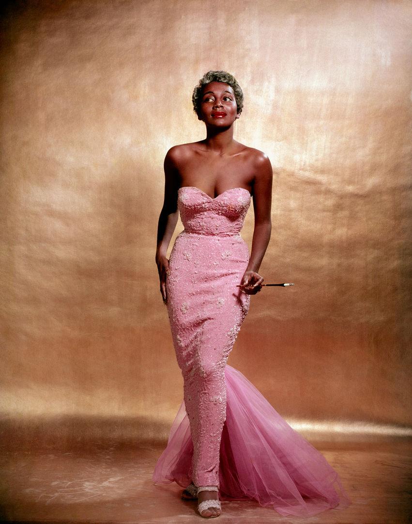 A woman standing up straight wearing a fitted pink dress. Her expression is entertaining, whole she hold her cigarette extension.