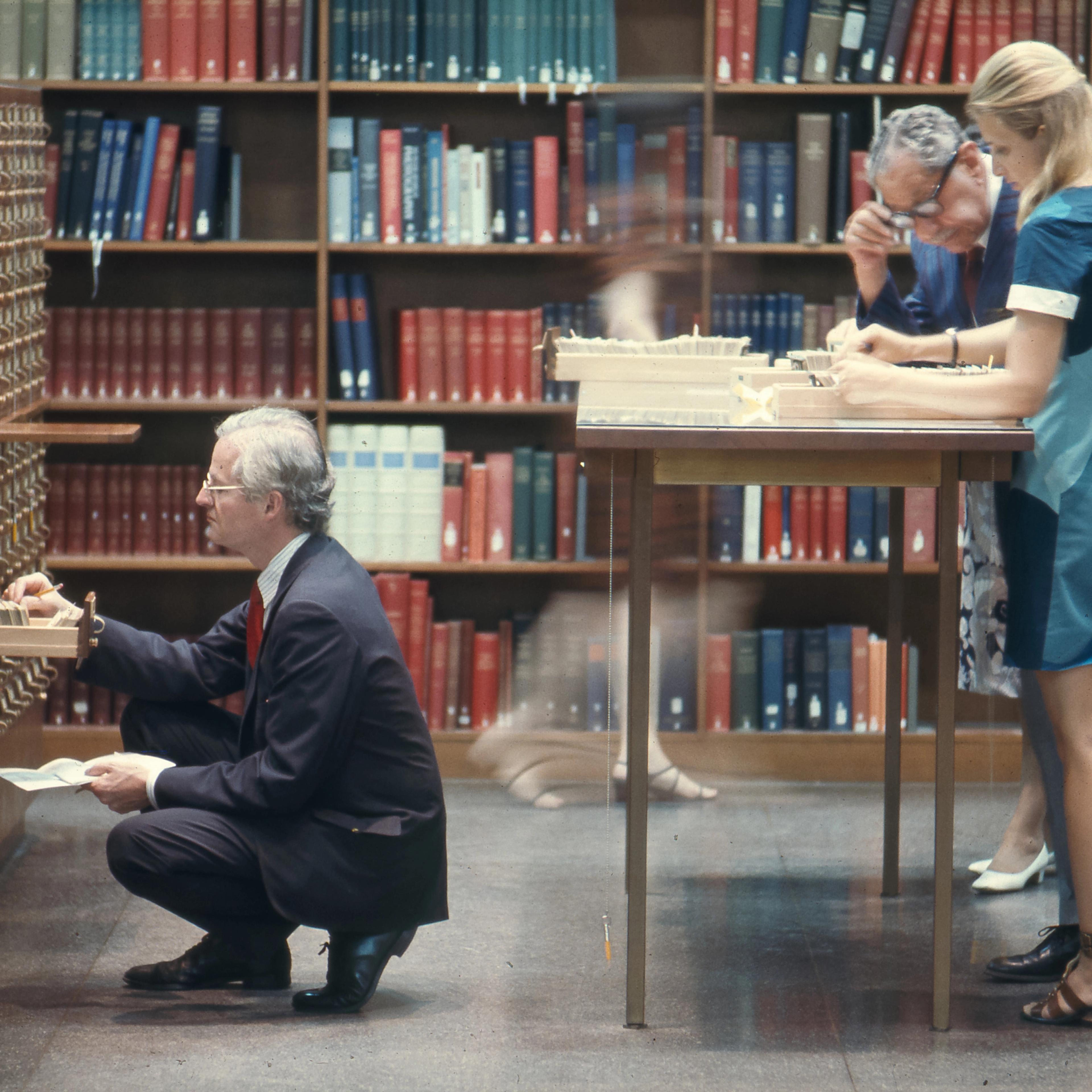 Dated photograph of people using a library card catalog and reference files, with bookshelves in the background.