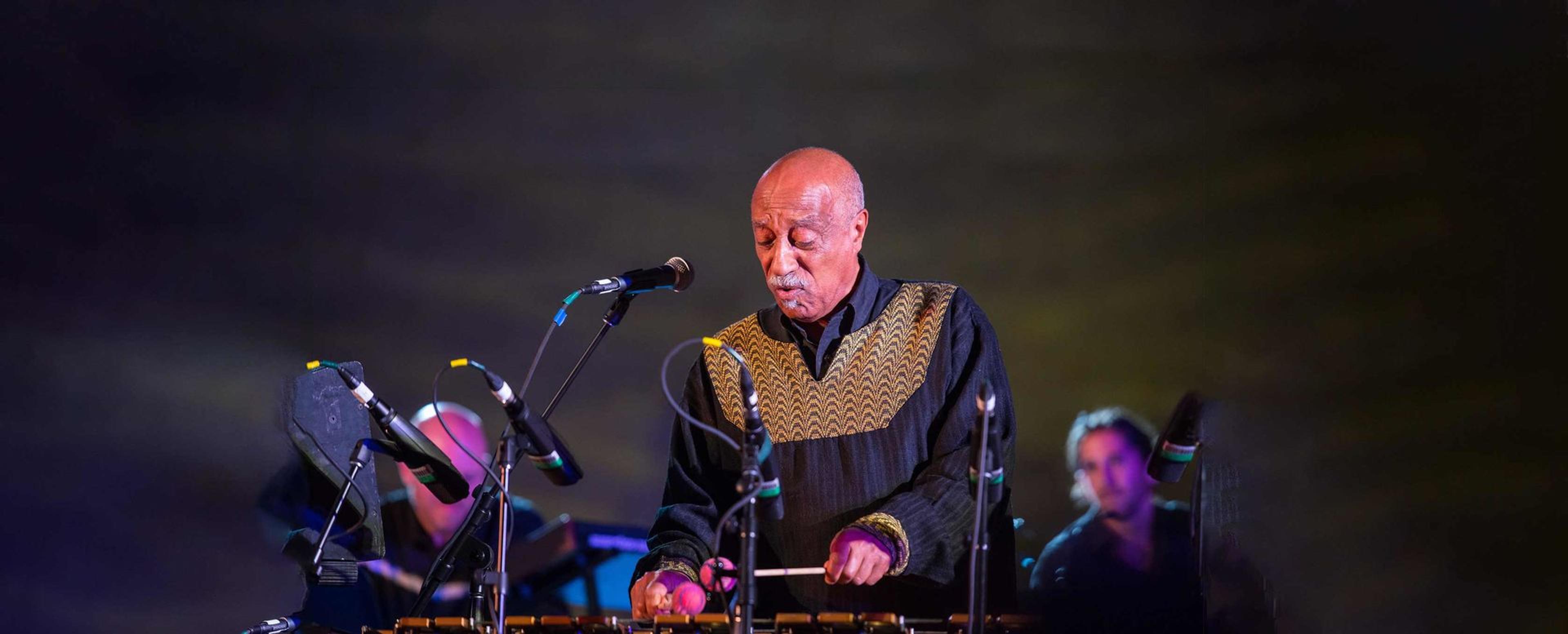 Mulatu Astatke plays the xylophone in front of a mic with other musicians in the backdrop.