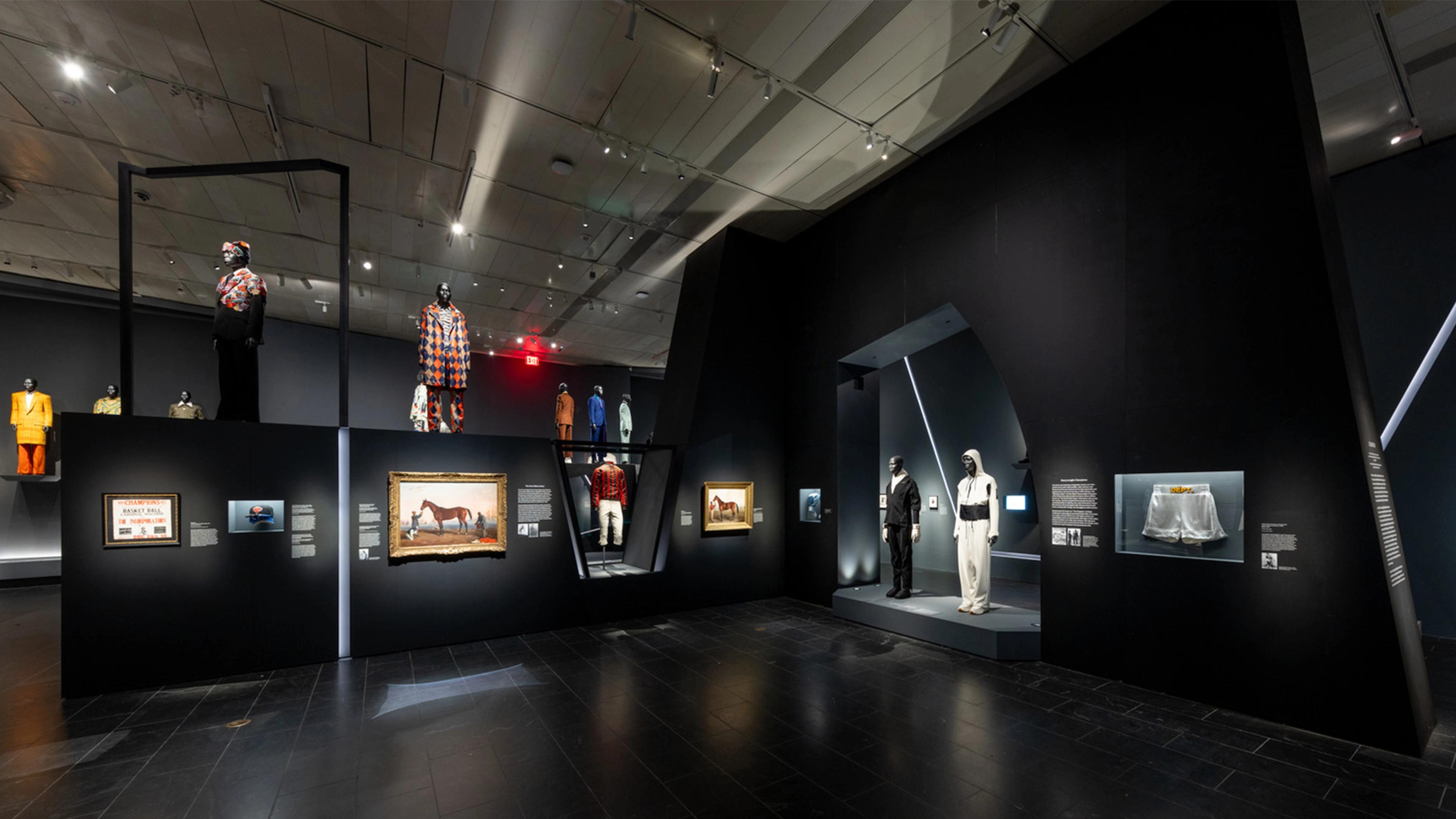Modern museum exhibit with mannequins wearing colorful, eclectic outfits and boxing shorts displayed in lit cases. Art and informative plaques adorn black walls.