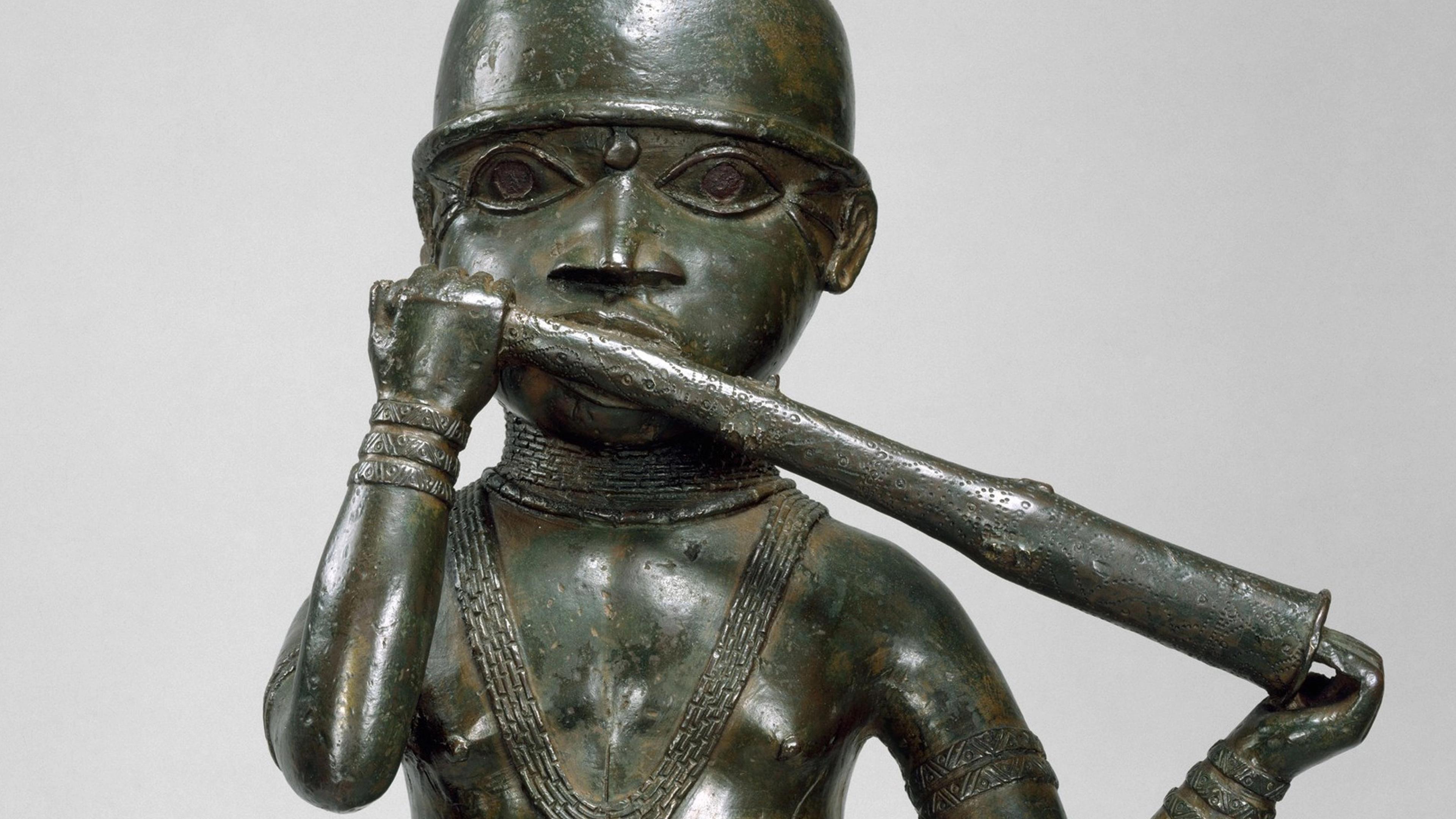 Bronze statue of a figure playing a long flute-like instrument, with stylized features and intricate jewelry.