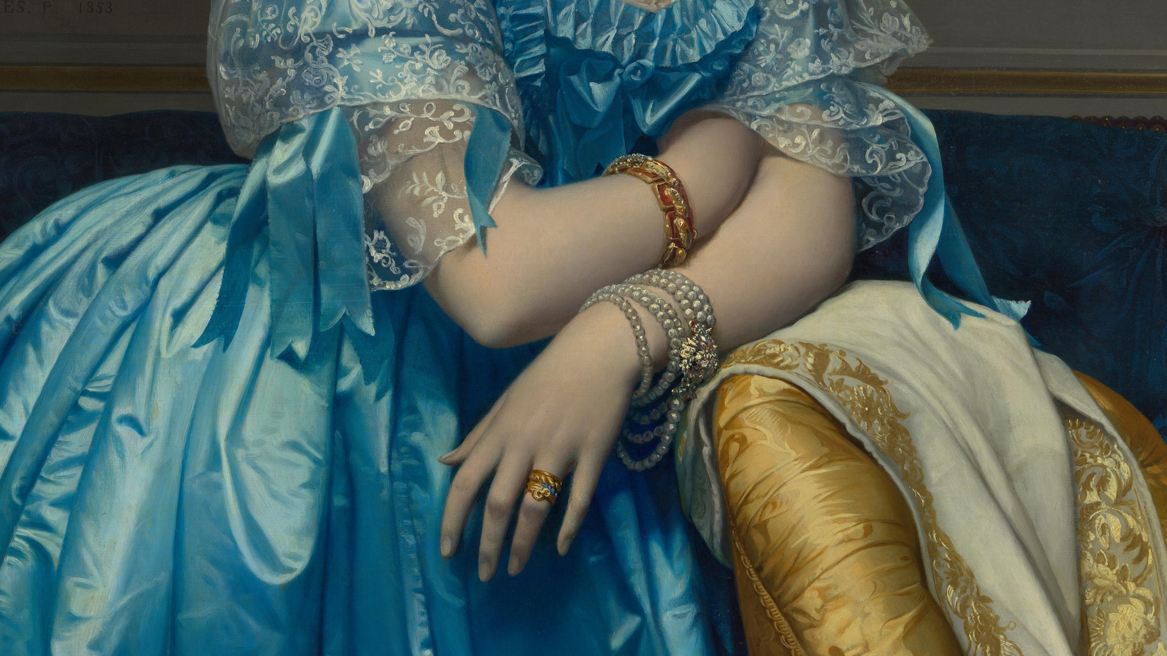 Close-up detail of a woman's hand resting against a yellow chair. She is wearing gold rings and a multi-stranded pearl bracelet.
