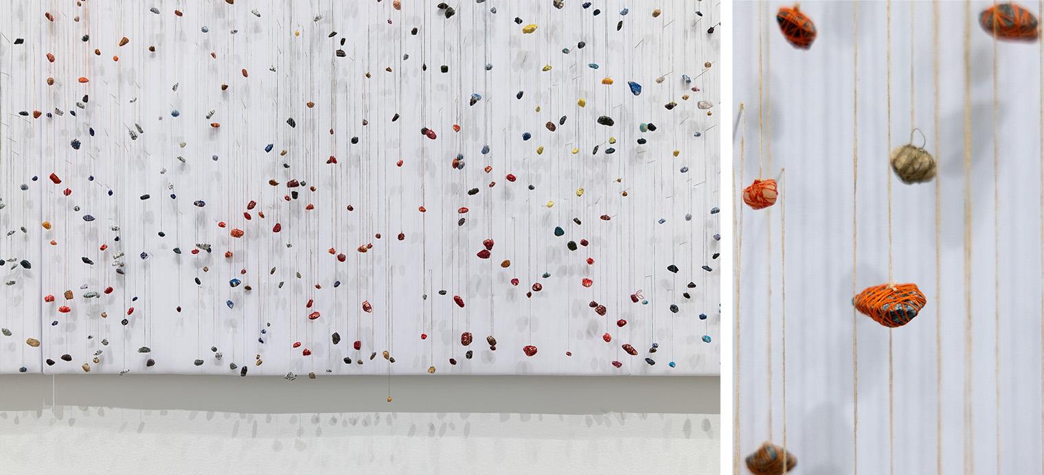Art installation featuring numerous rocks suspended on vertical strings against a white background. A close-up showcases orange thread-wrapped rocks.