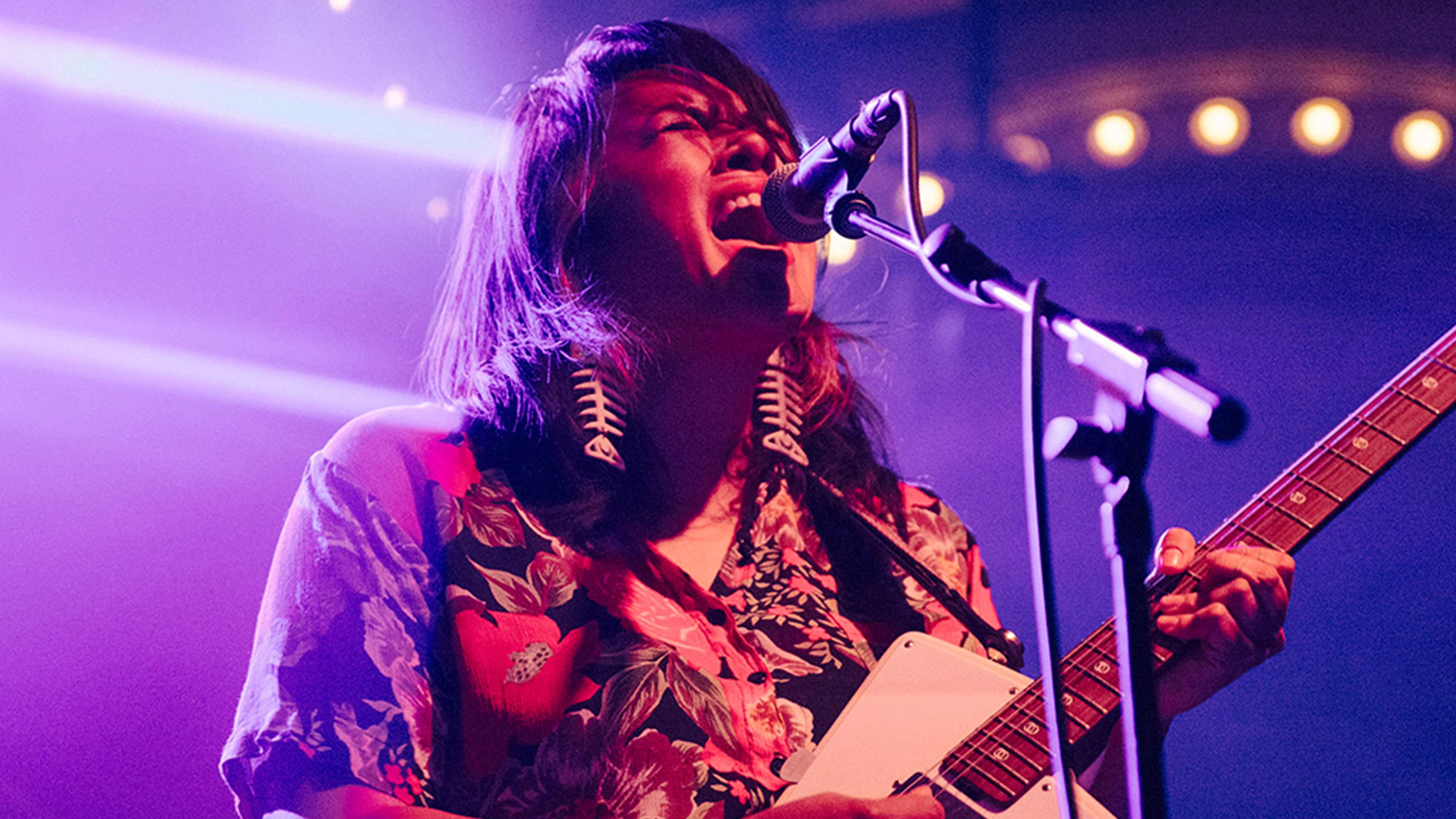 A long-haired musician sings passionately into a microphone while playing a white electric guitar onstage, bathed in vivid purple and red lights.