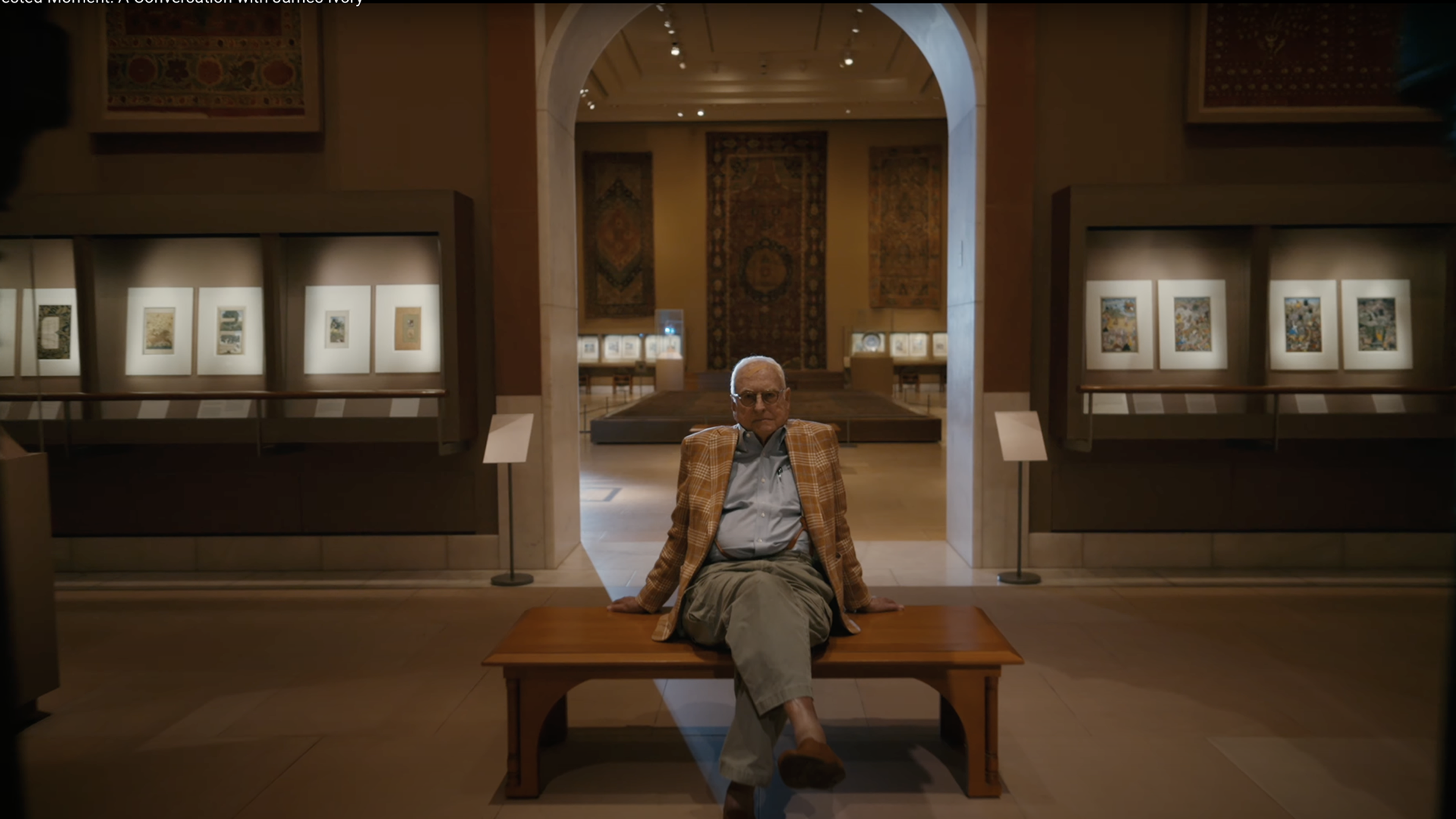 Image of James Ivory sitting on a bench the gallery "Later South Asia", with paintings and carpets in the background.