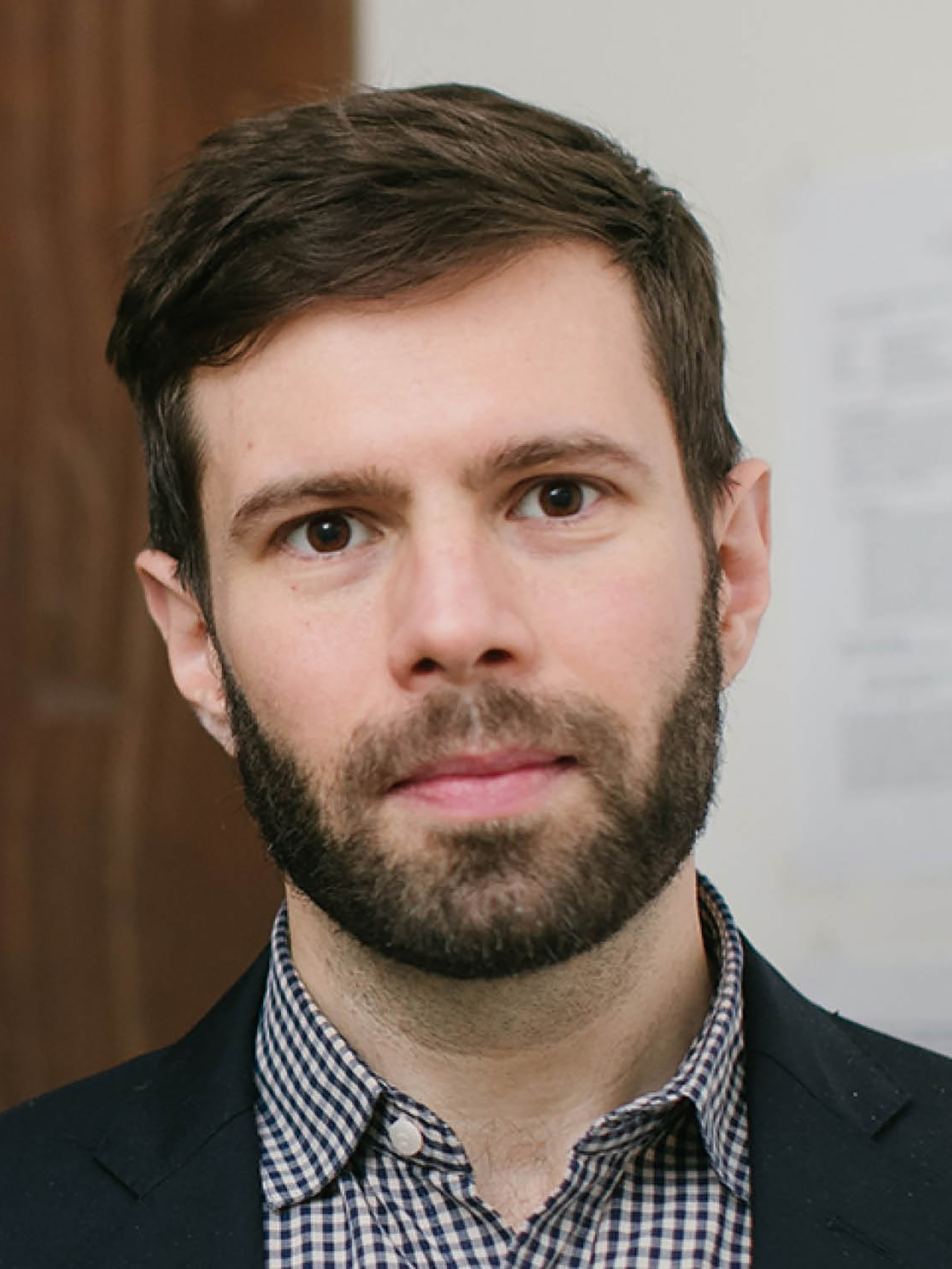 A bearded man wearing a plaid shirt and blazer stands against a neutral background