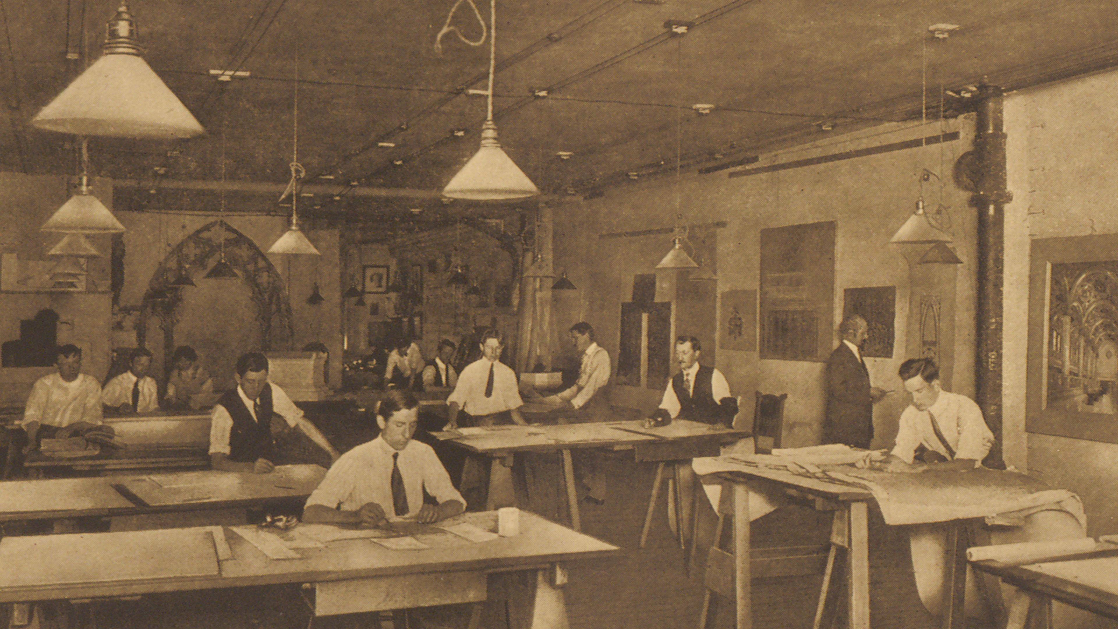 Sepia photograph of men in suits working quietly at large drafting tables in a vintage office, beneath hanging lamps