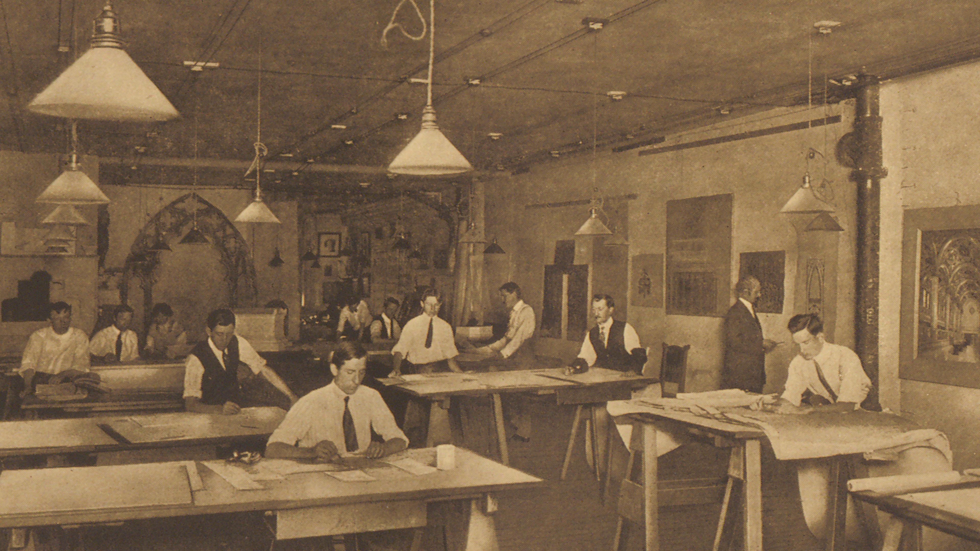 Sepia photograph of men in suits working quietly at large drafting tables in a vintage office, beneath hanging lamps