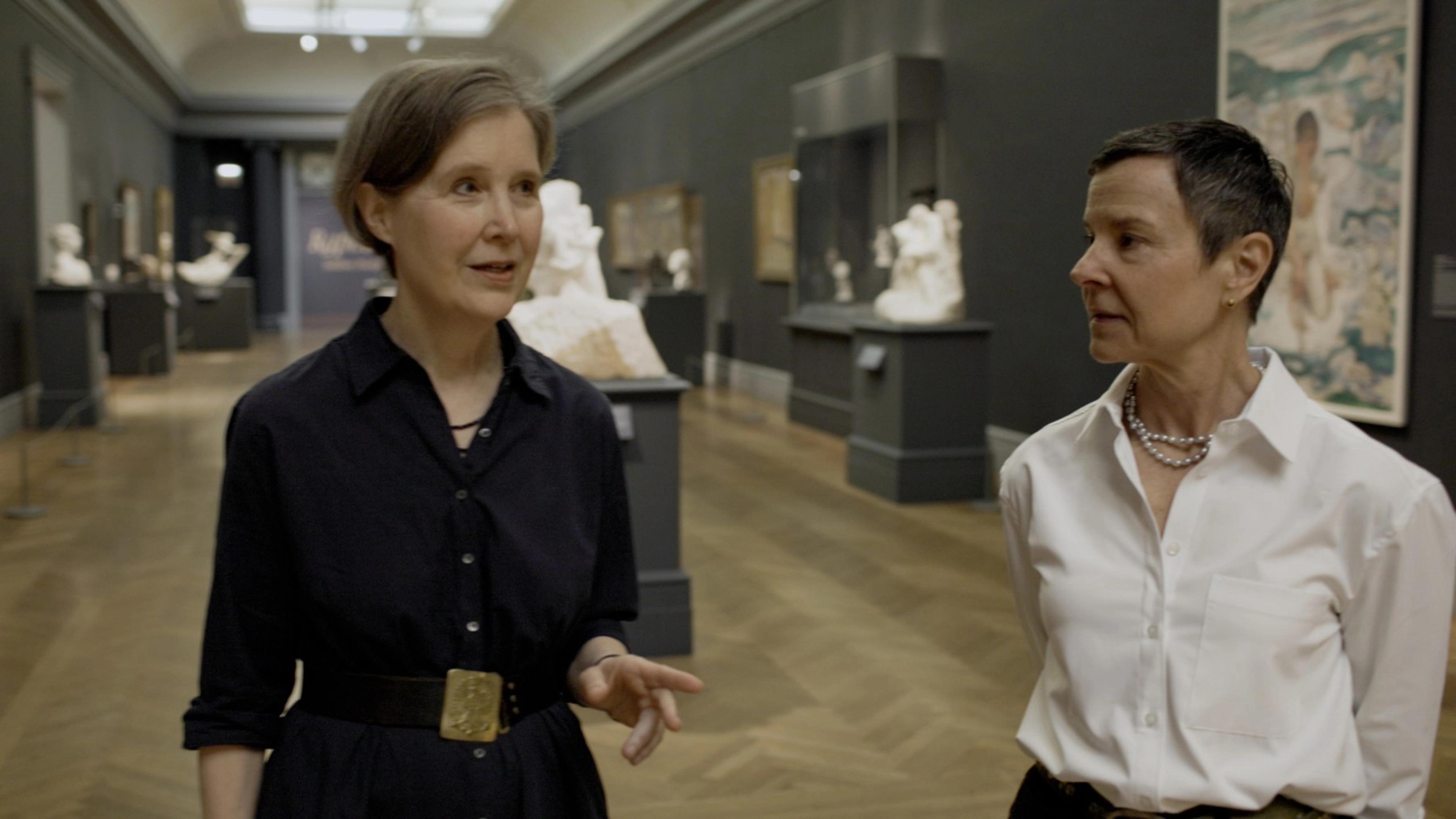 Two women angled towards each other in a skylit gallery with white statues on pedestals in the background.
