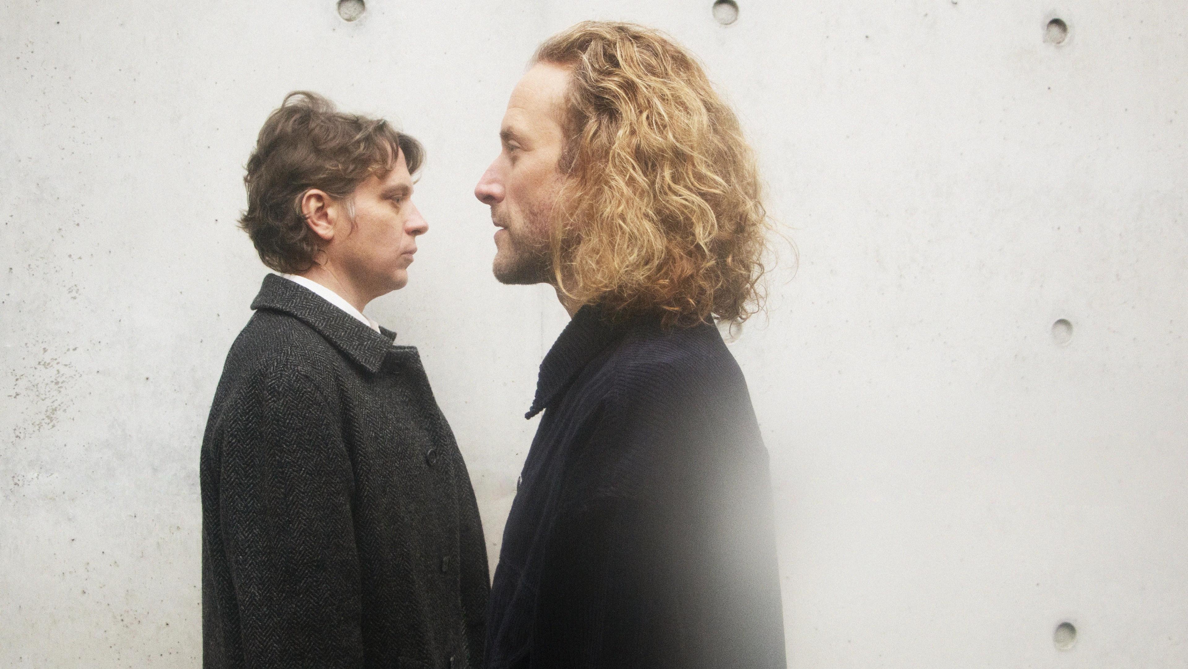 In the foreground, a man with blond curly hair; in the background, a man with short brown hair. They both wear trench coats and face each other to the side.