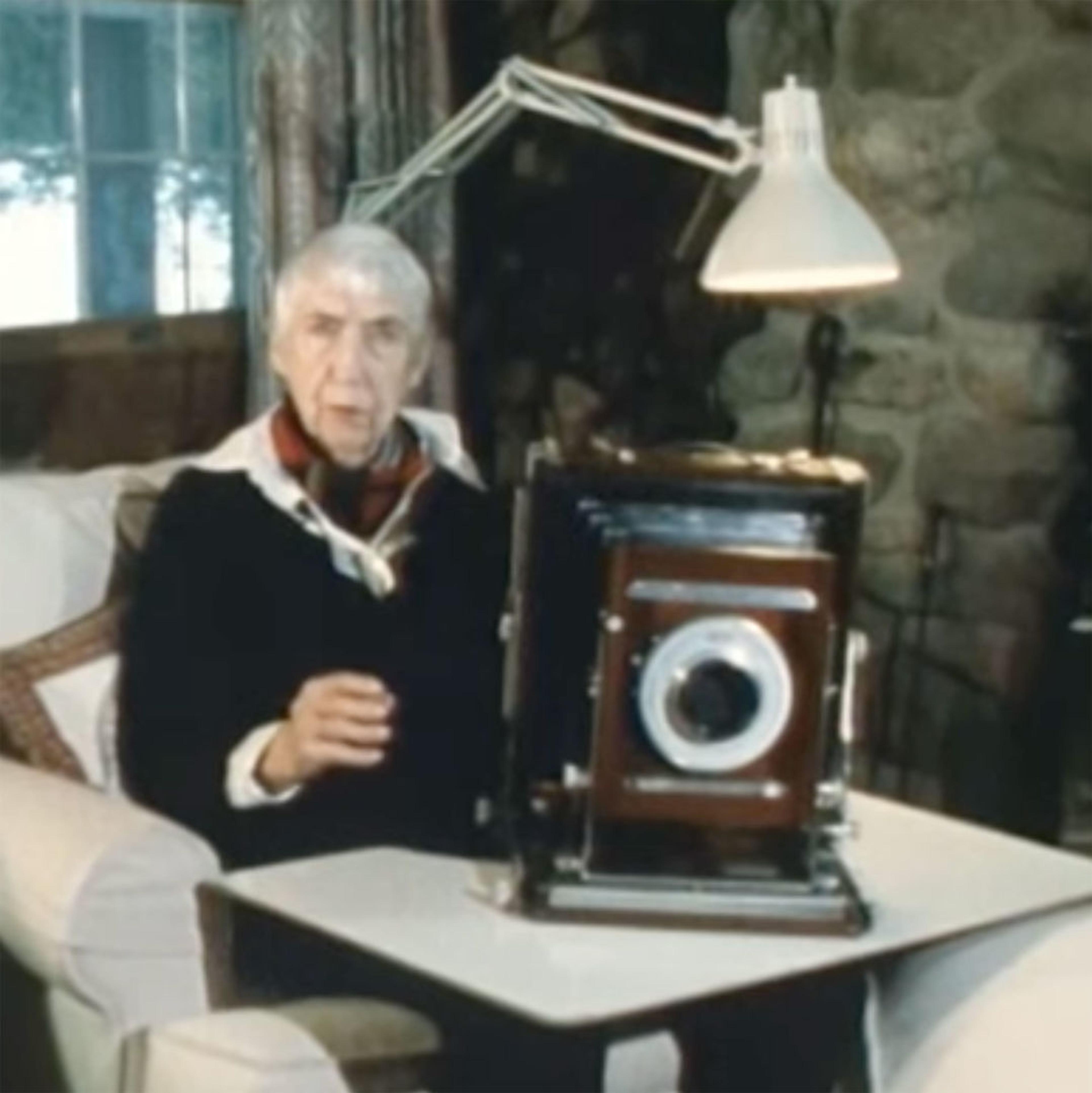 Still from "The Living Room" of Berenice Abbott sitting in an armchair in front of a fire with a camera on the table in front of her