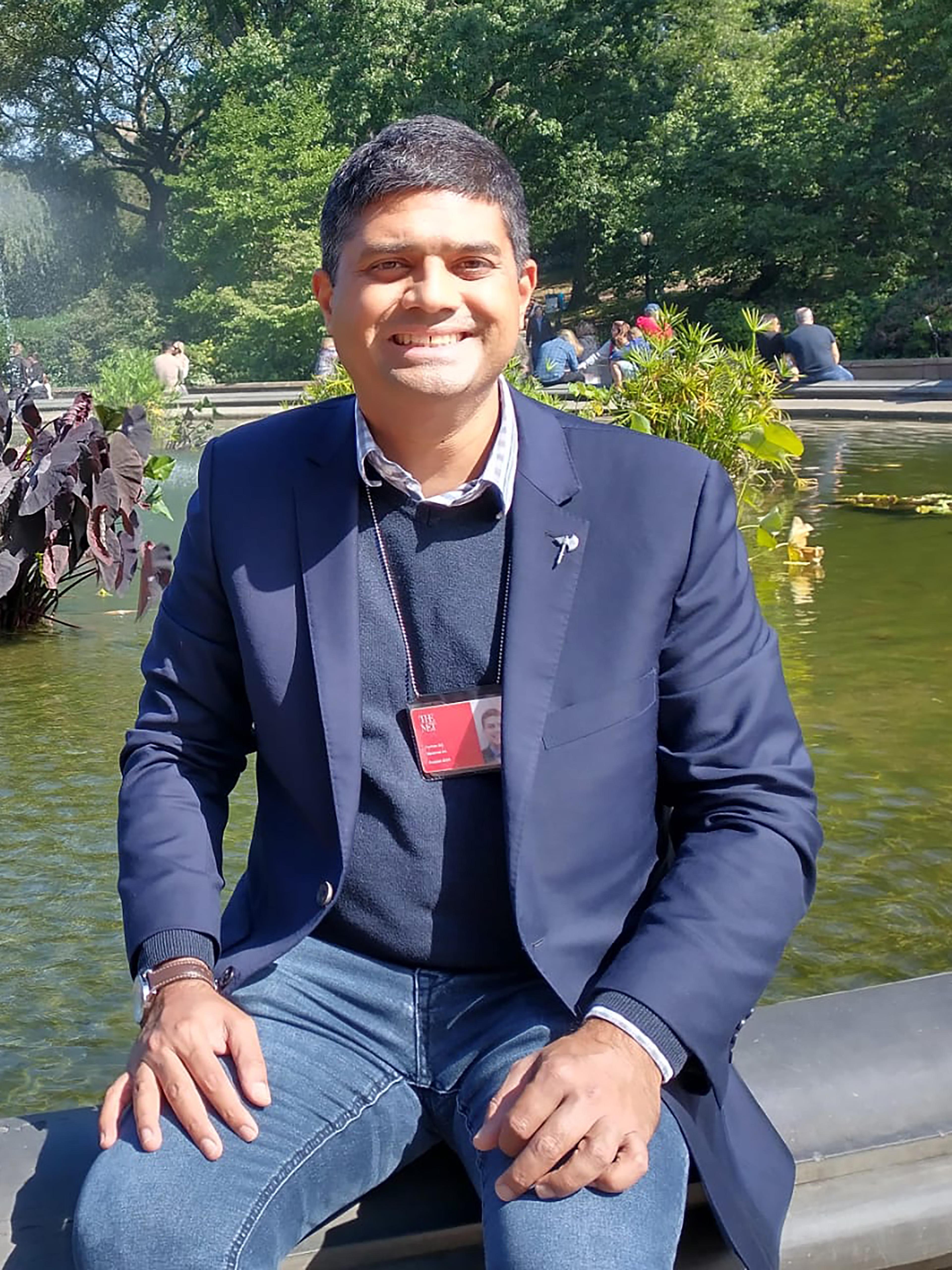 A man wearing a navy blazer sits on the edge of a fountain and smiles