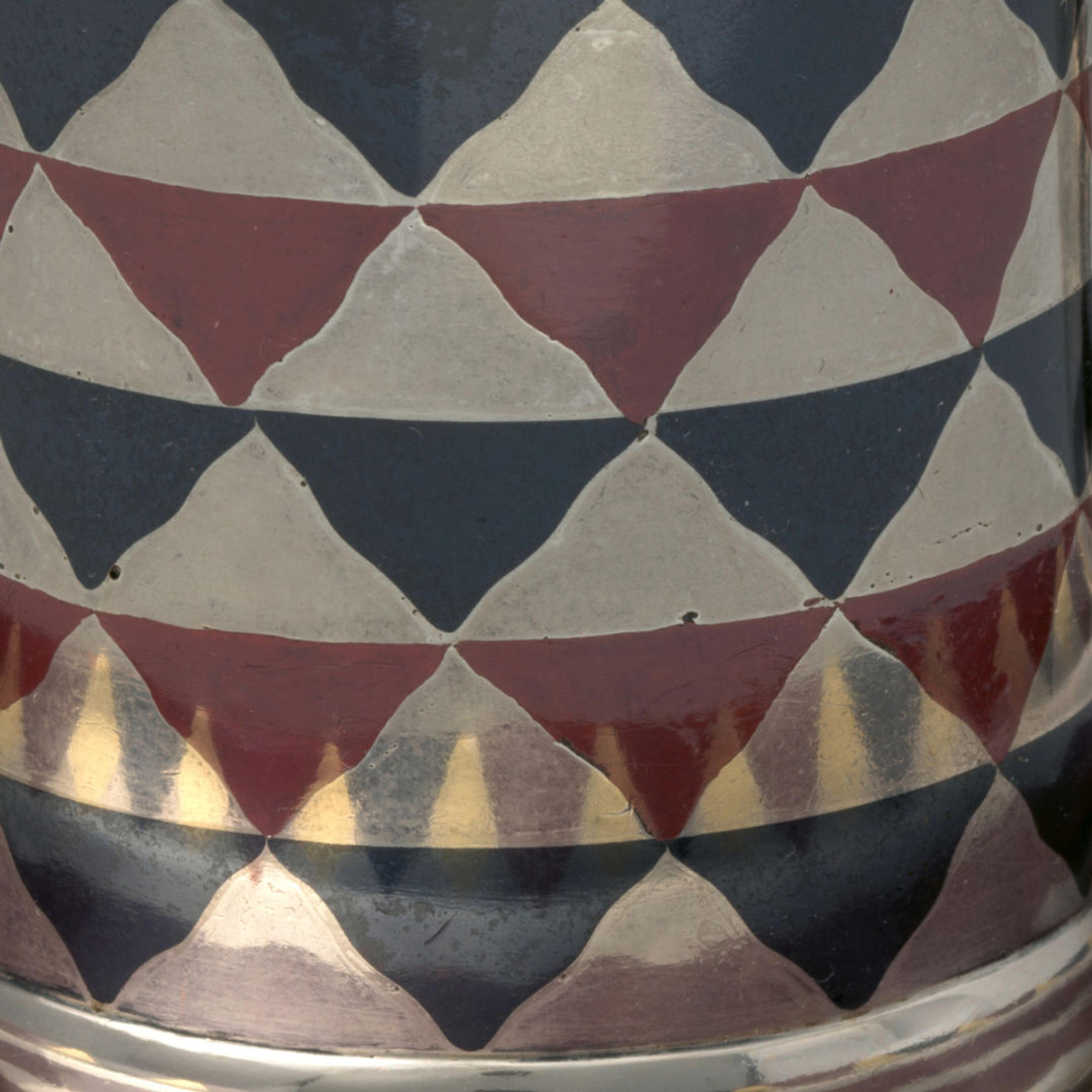 A silver, blue, and maroon cylindrical cup with a handle rests on a matching saucer. The cup and saucer are reflective and adorned with an inlay design of a checkerboard pattern of wavy triangle shapes.
