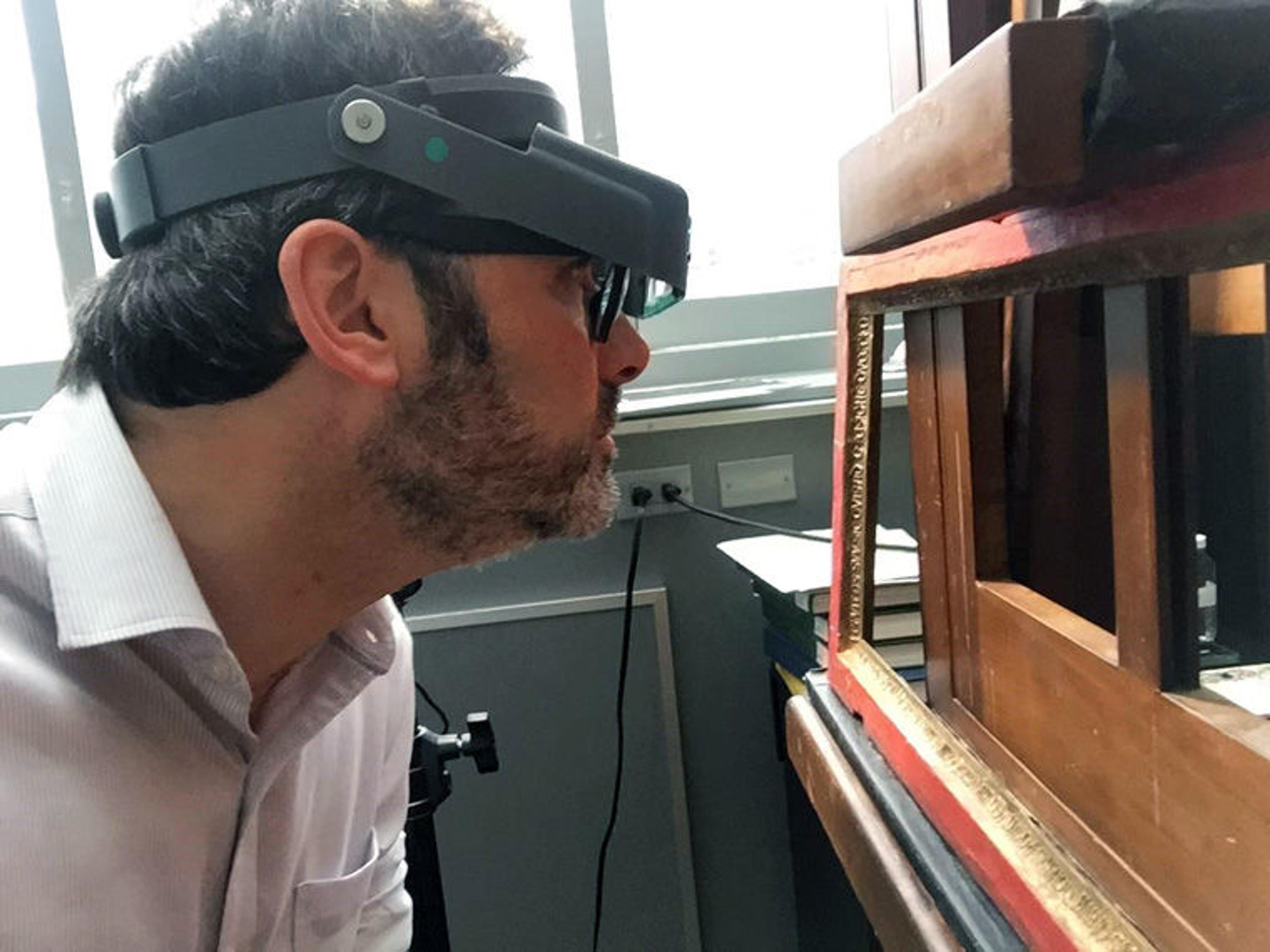 A man wearing a headpiece with magnifying glass closely inspects a wooden painting frame in a sunlit conservation studio