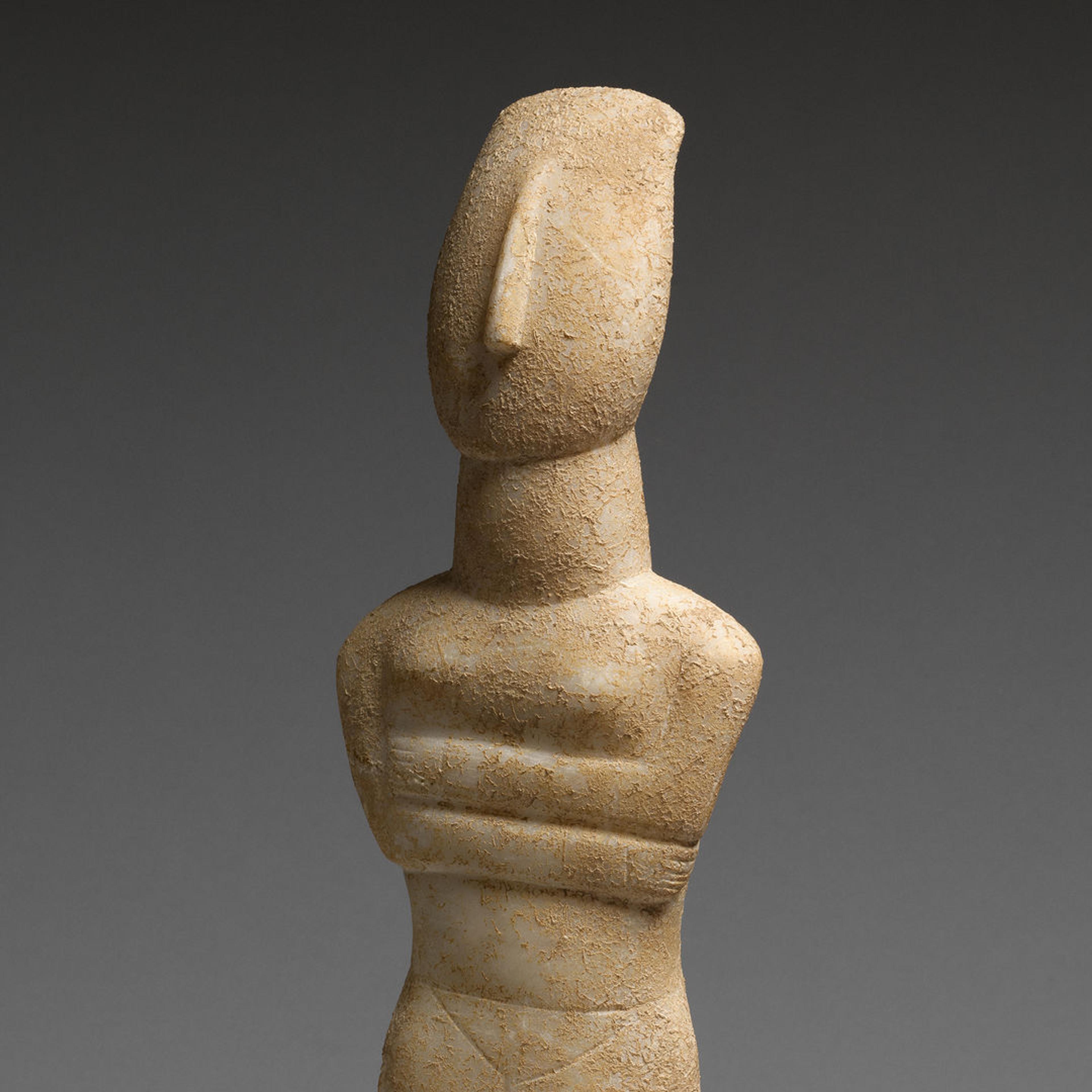 Nude stone Cycladic figure with it's arms crossed against a grey background.