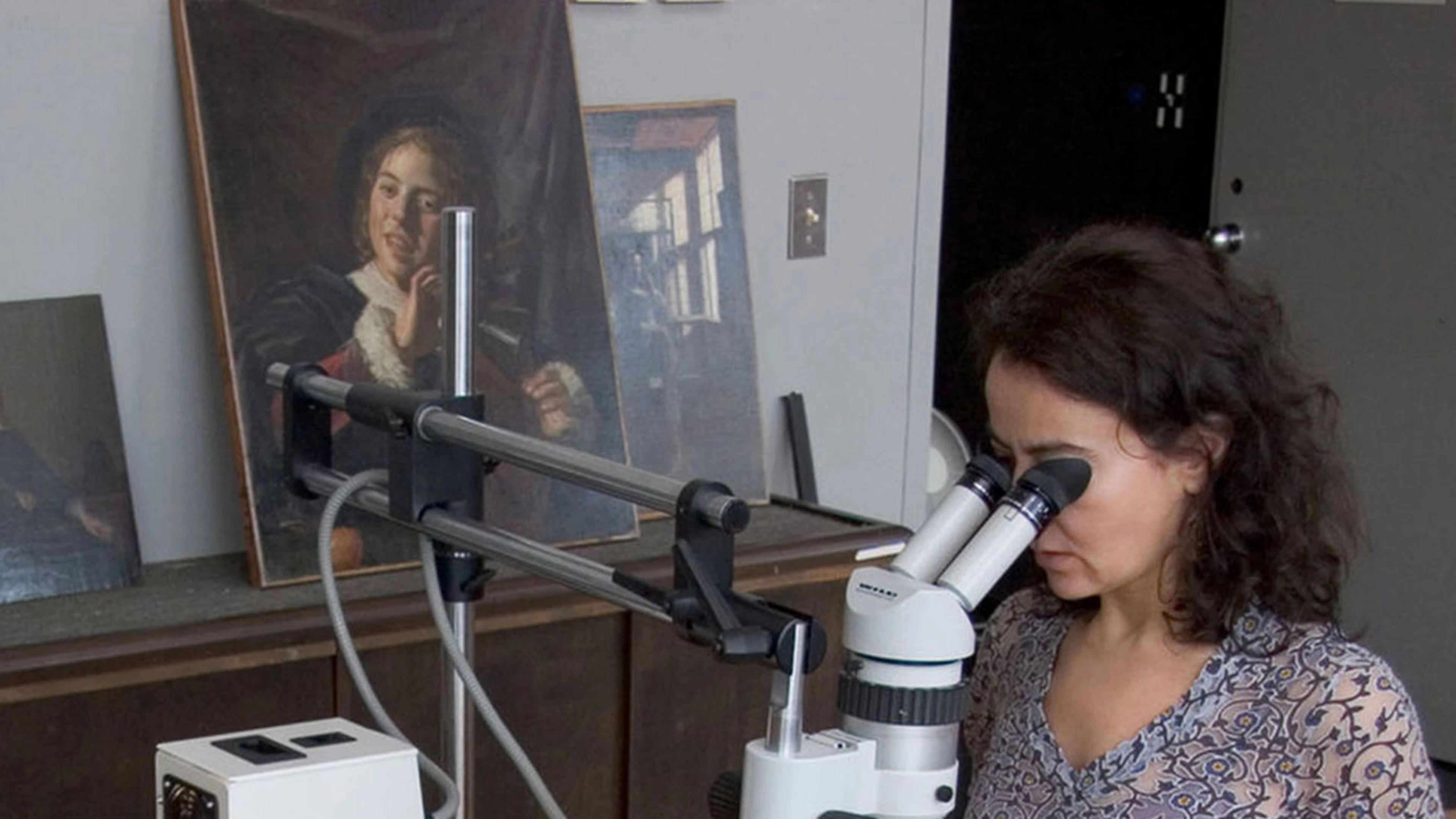 A woman examines art through a microscope, surrounded by framed paintings on a tabletop in a studio setting.