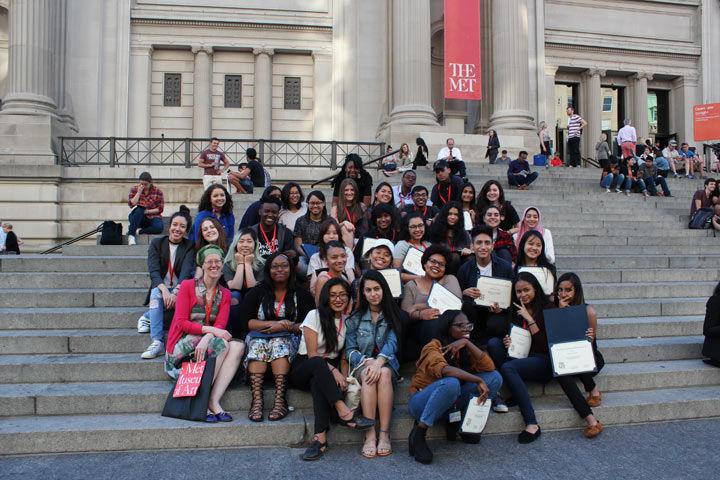 A large group of young interns sit on the steps in front of the Museum with their certificates for completing the internship