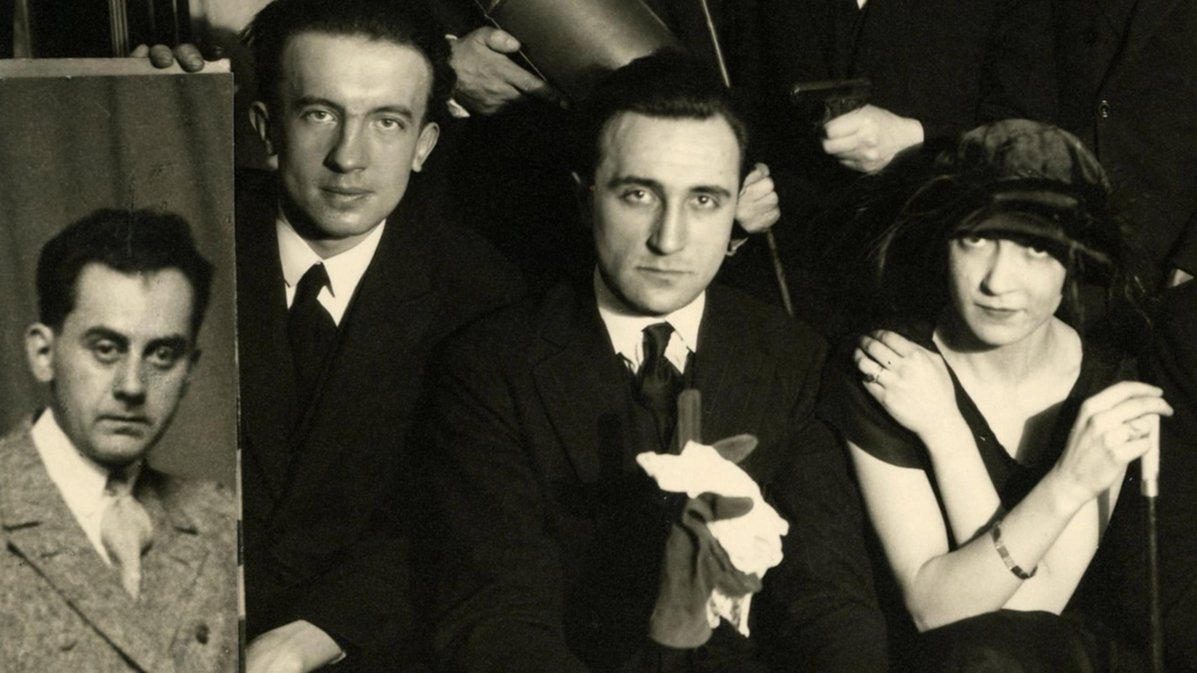 Black-and-white photograph of two men and one woman. The man on the left holds up a life-size portrait of another man.