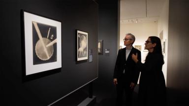 A man and woman in a dimly lit gallery observe black-and-white abstract art on dark walls. The mood is focused and contemplative.