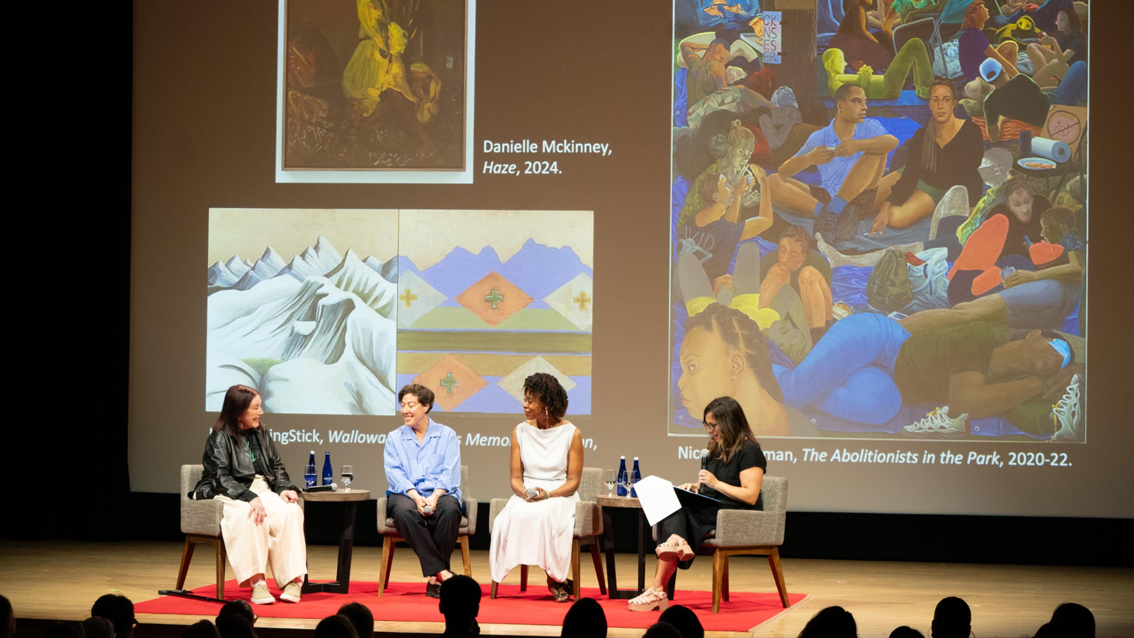 Four women in conversation on a stage. A slide with large, colorful artworks is projected behind them.