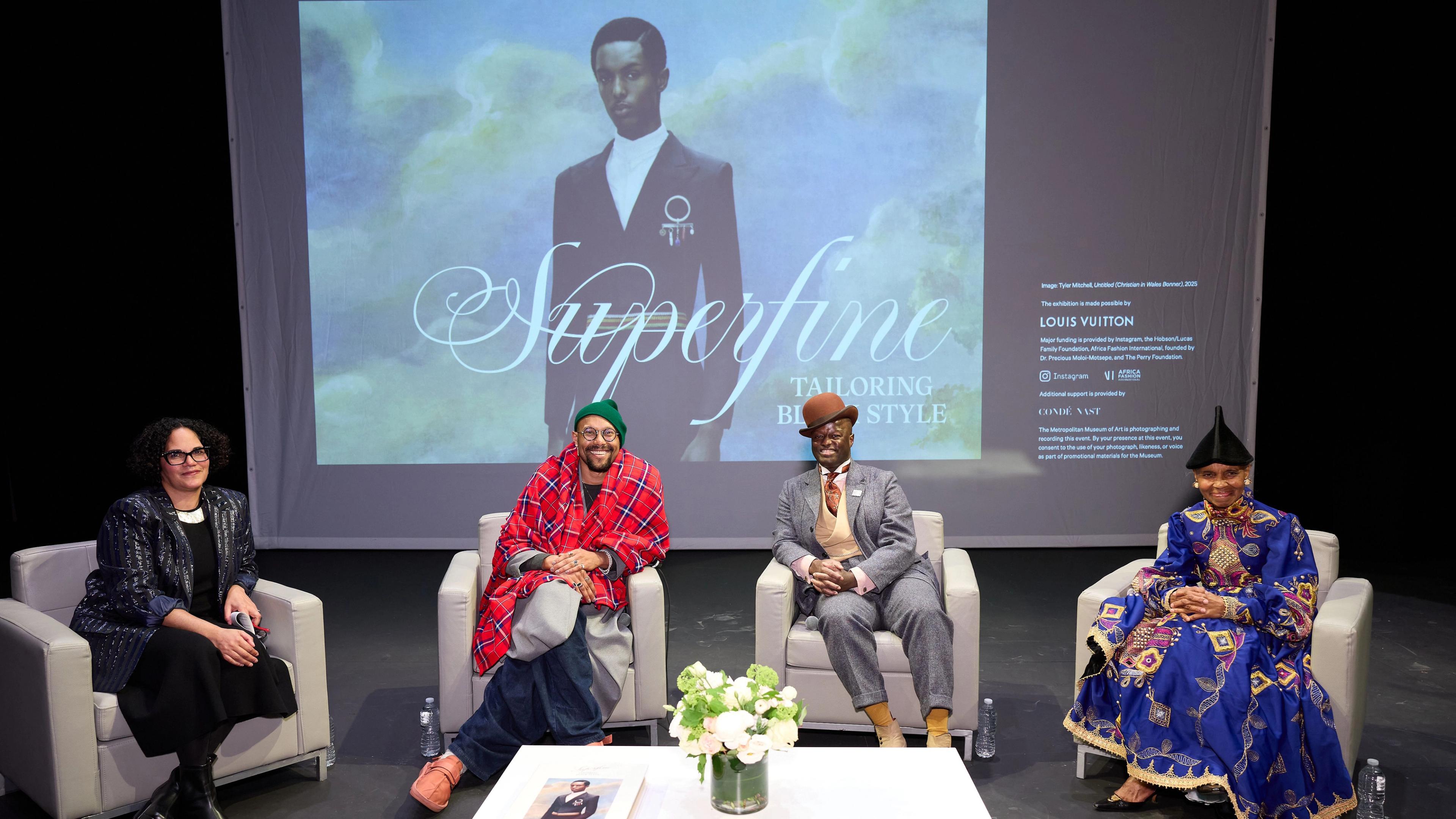 Four individuals are seated on stage in a panel discussion. Behind them, a screen shows "Superfine Tailoring Black Style" and an elegant fashion image. The setting appears formal and thoughtful.