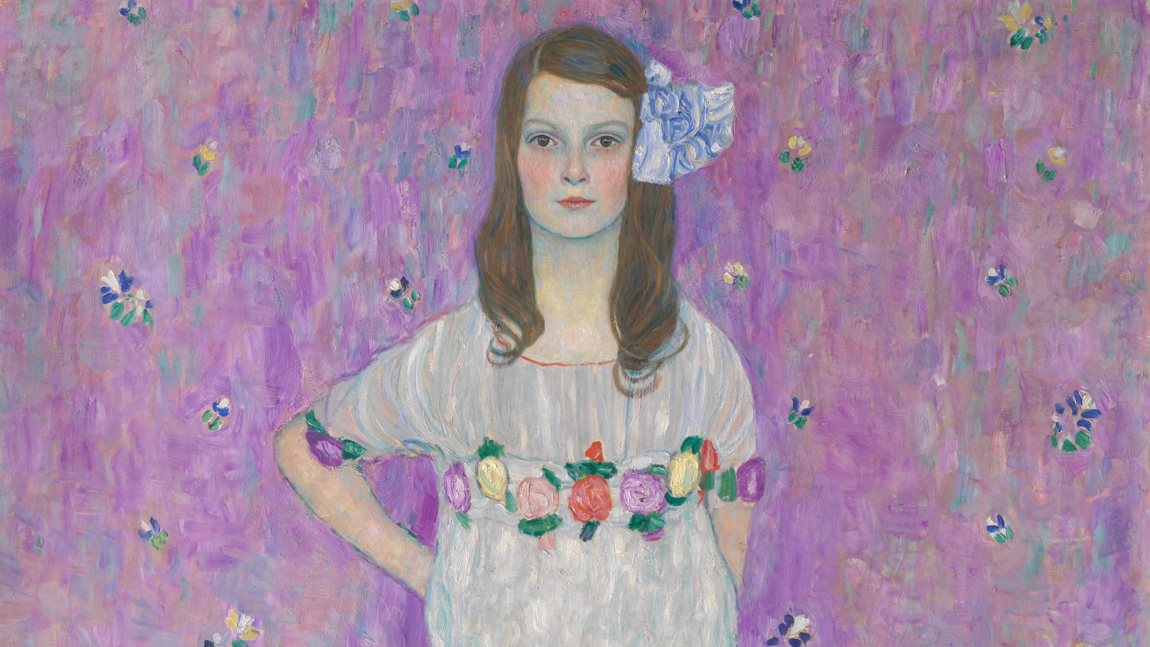 A young girl poses with a stoic face and a hand on her hip. Her dress and hair are accented with flowers.