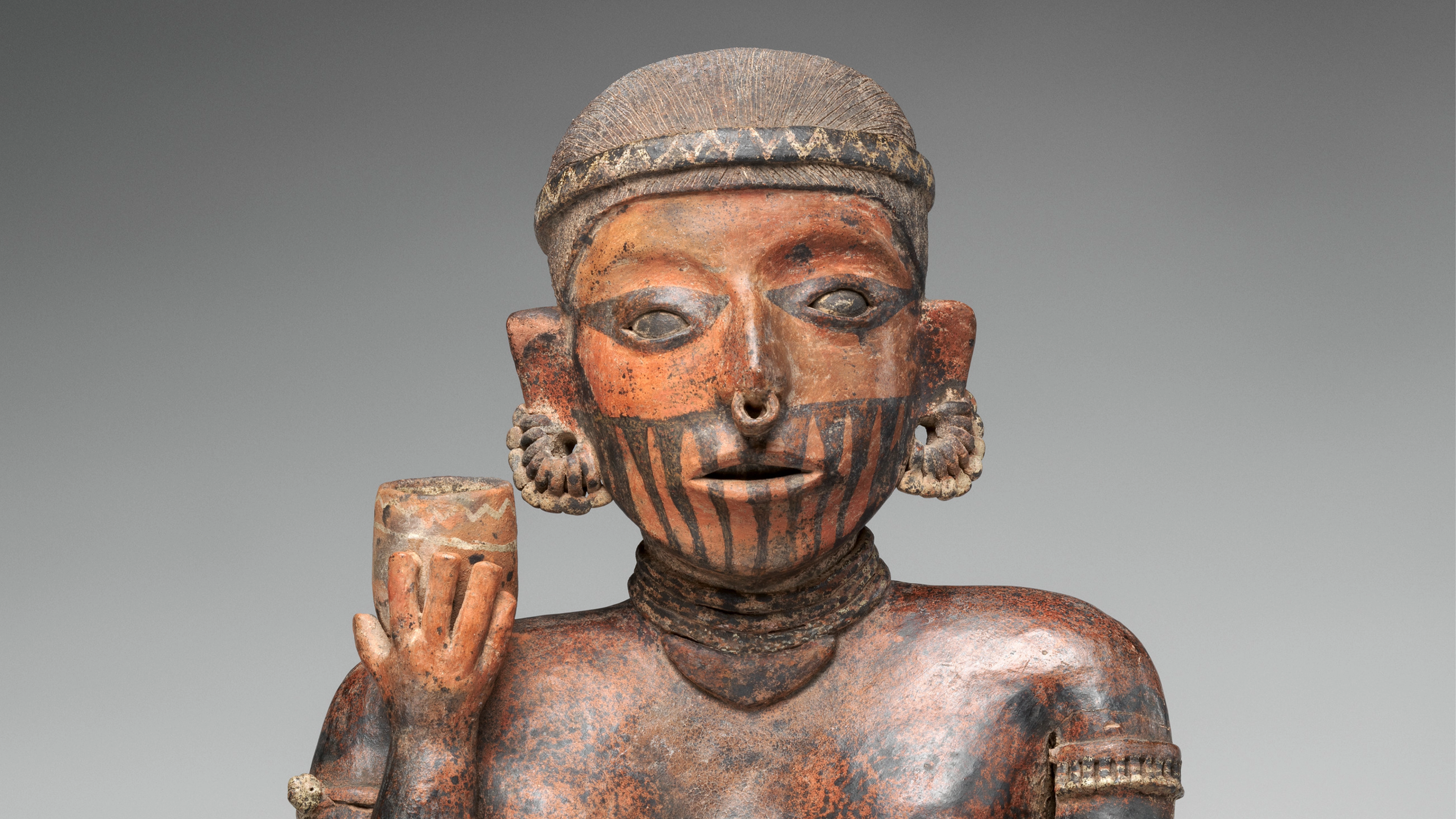 Ceramic female figure adorned with jewelry and face markings, holding up a cup in one hand.