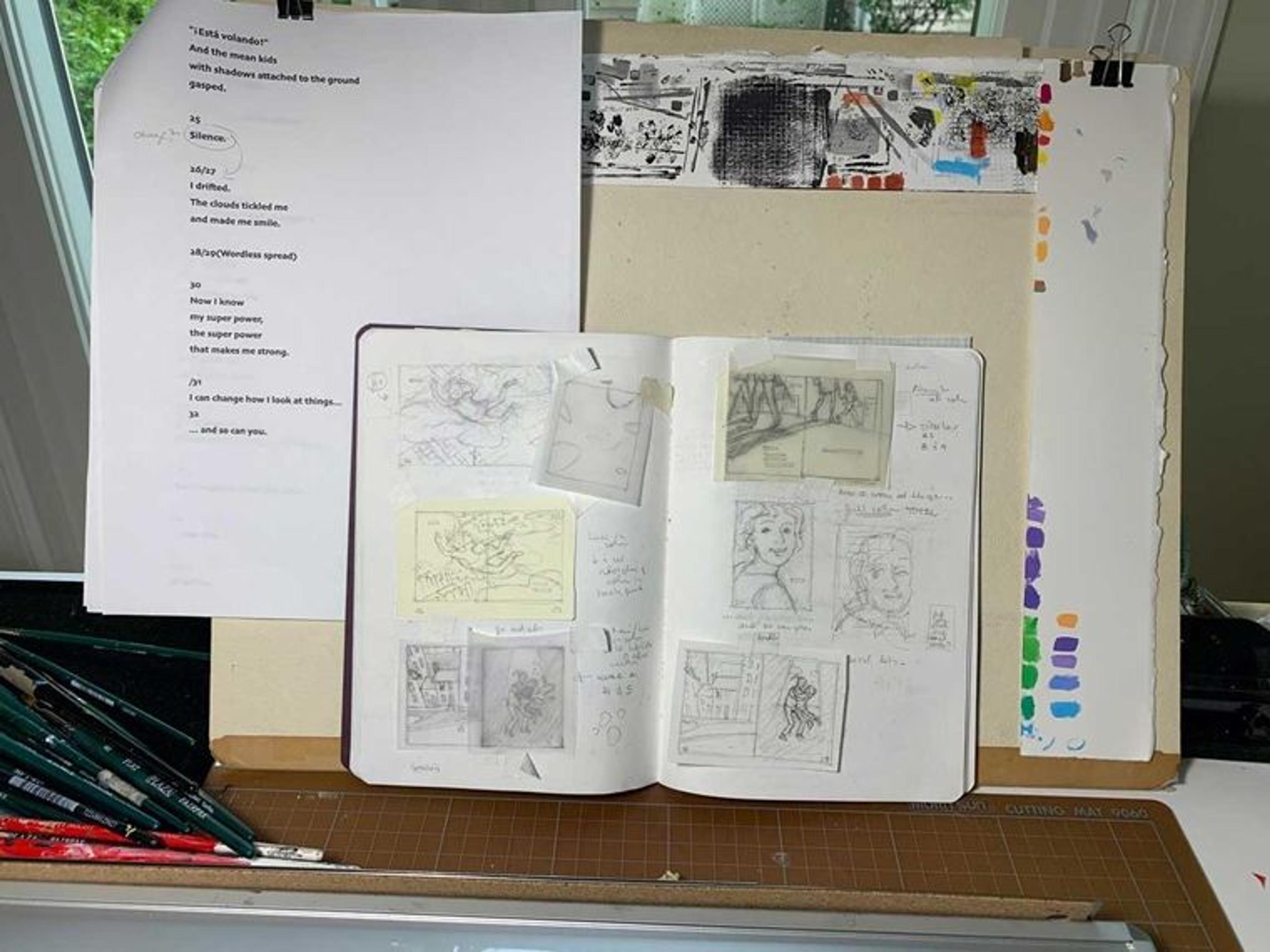 An open sketchbook sits on top of an artist's workbench. Small drawings of illustrations from the book Luci Soars are drawn in pencil on the open pages. under the notebook is a printout of text that will later accompany the illustrations in the book.