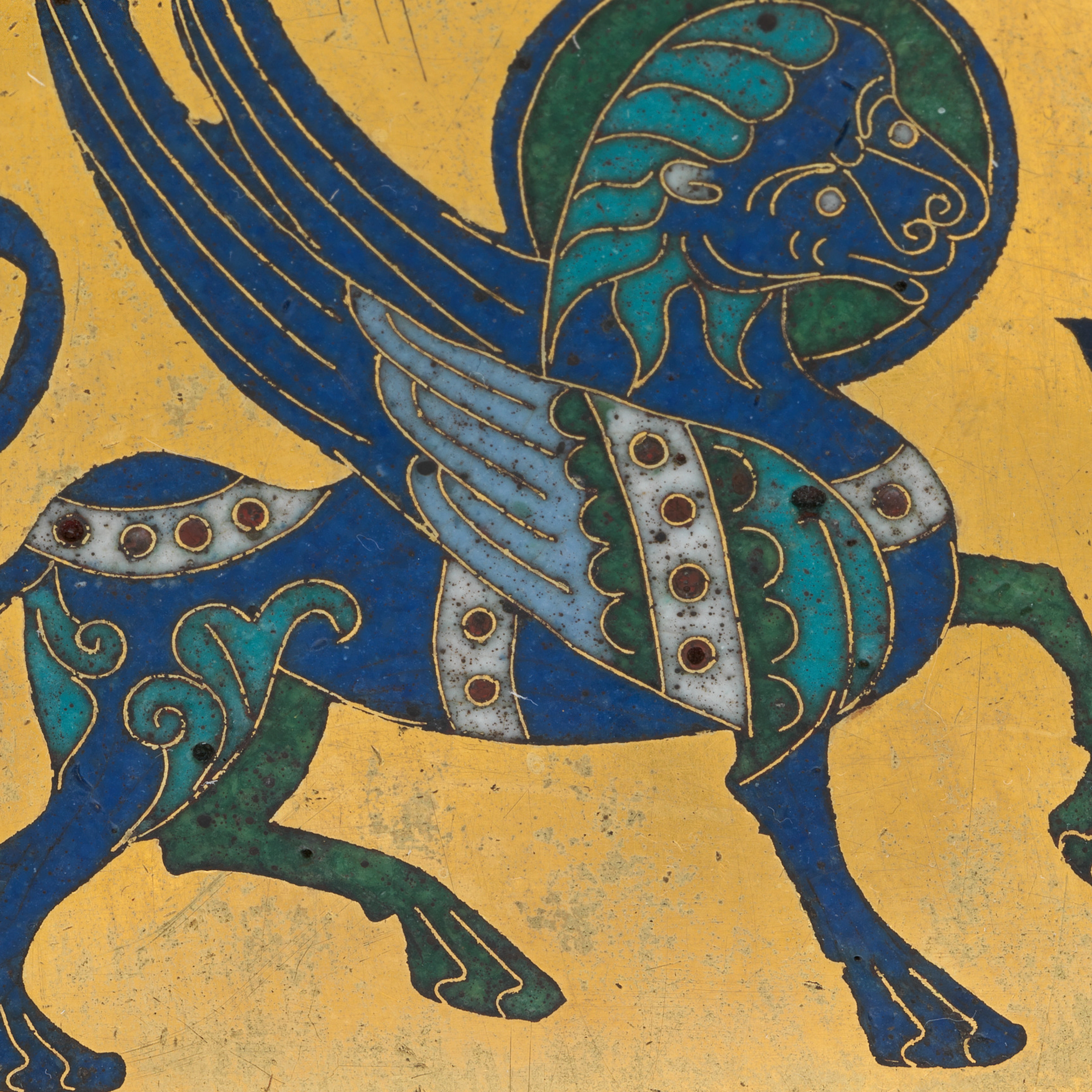 An ornate blue mythical creature with sweeping wings, curled tail, and patterned feathers fills a worn gold manuscript page beside dark decorative letters.