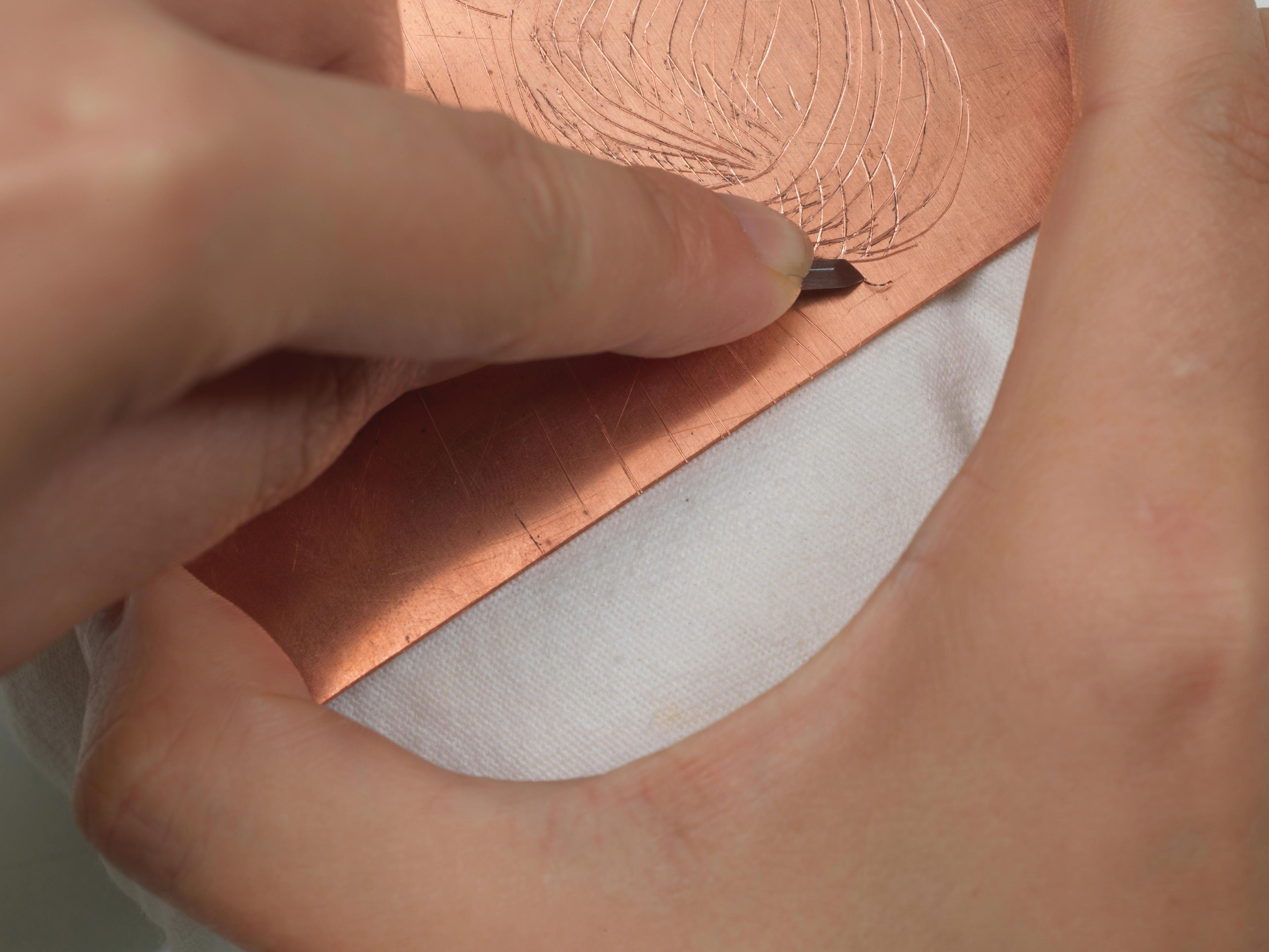 Close-up image of a printmaker's hands using a burin to incise a copper engraving plate.