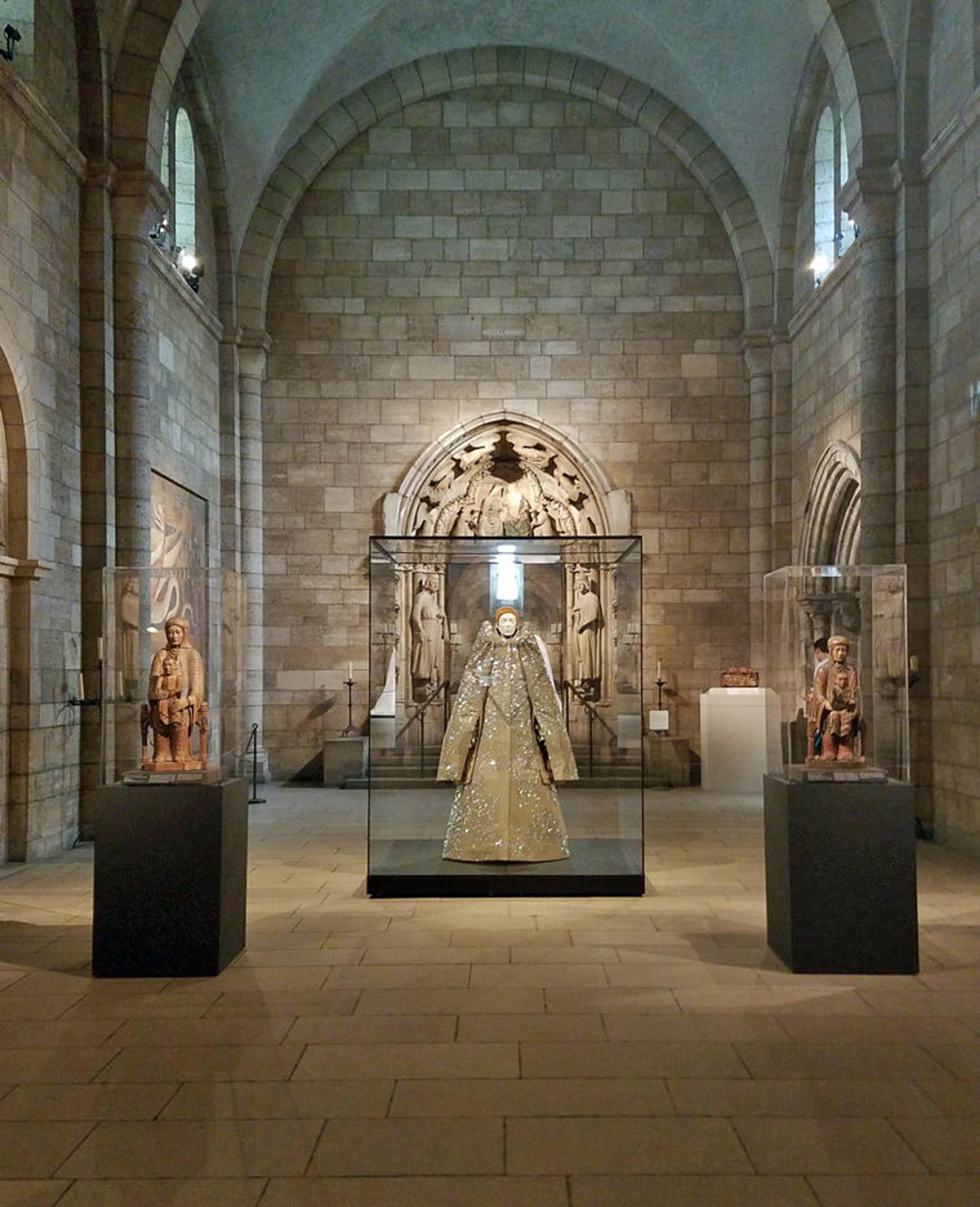 View of a gallery in a medieval cloister, featuring two twelfth-century sculptures of the Virgin and Child on either side of a contemporary ensemble by Viktor & Rolf