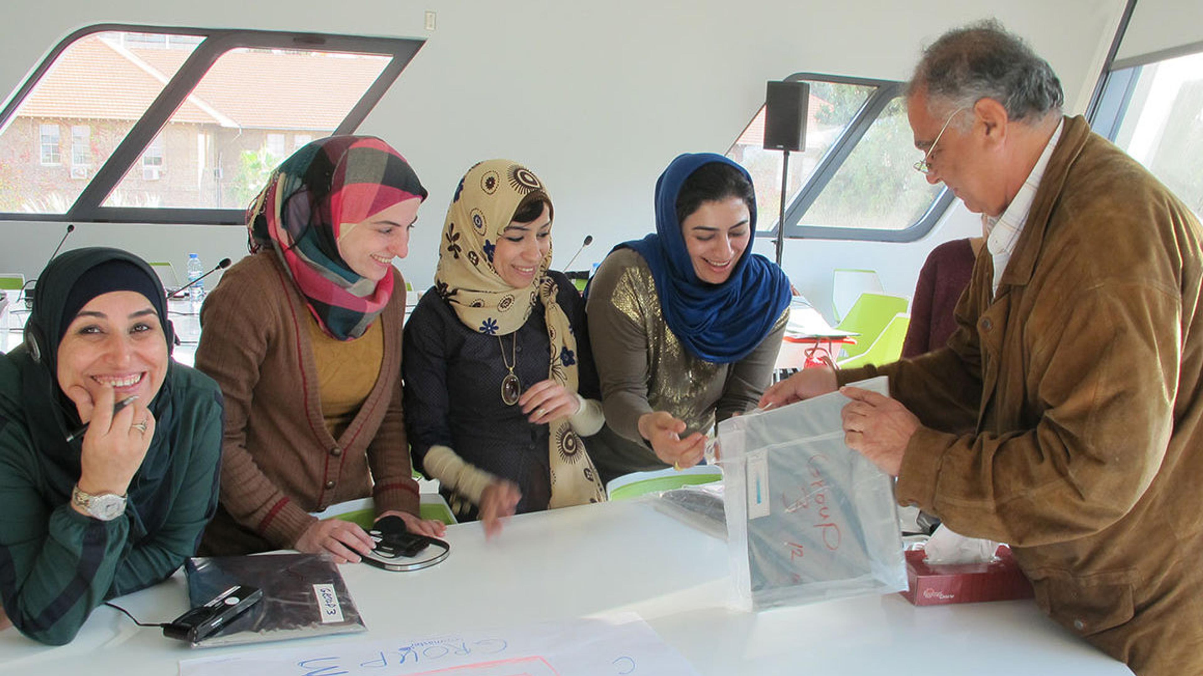Four young women on left side of work table, watching as male instructor to the right of the table demonstrate an enclosure for photographs.