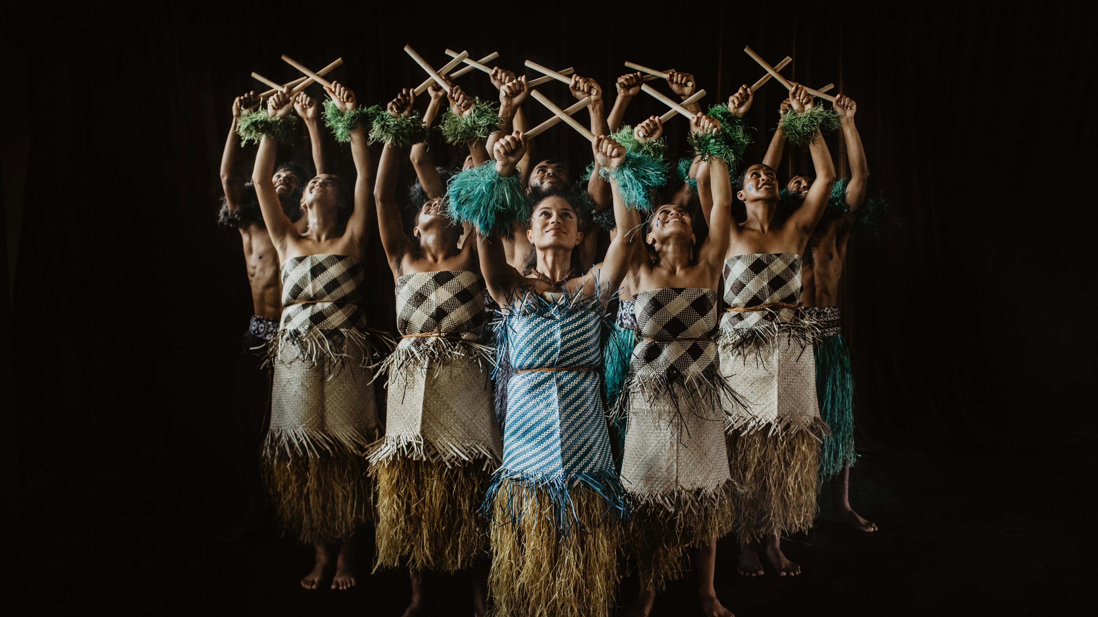 A group of performers in wrapped clothing hold sticks crossed in the air