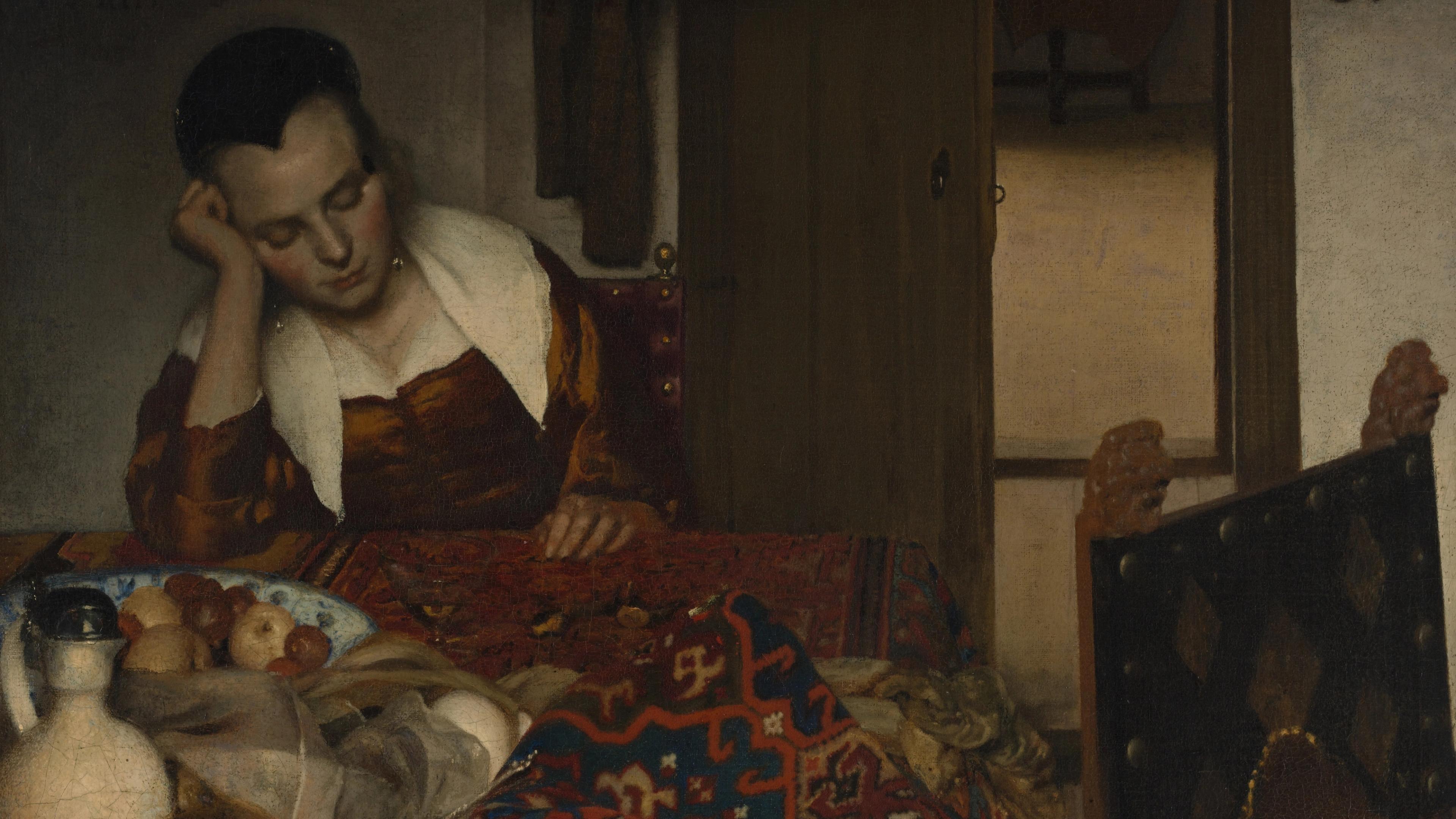 A woman rests against her hand propped up on a table. The scene features a bowl of fruit, a jug, and patterned prints.