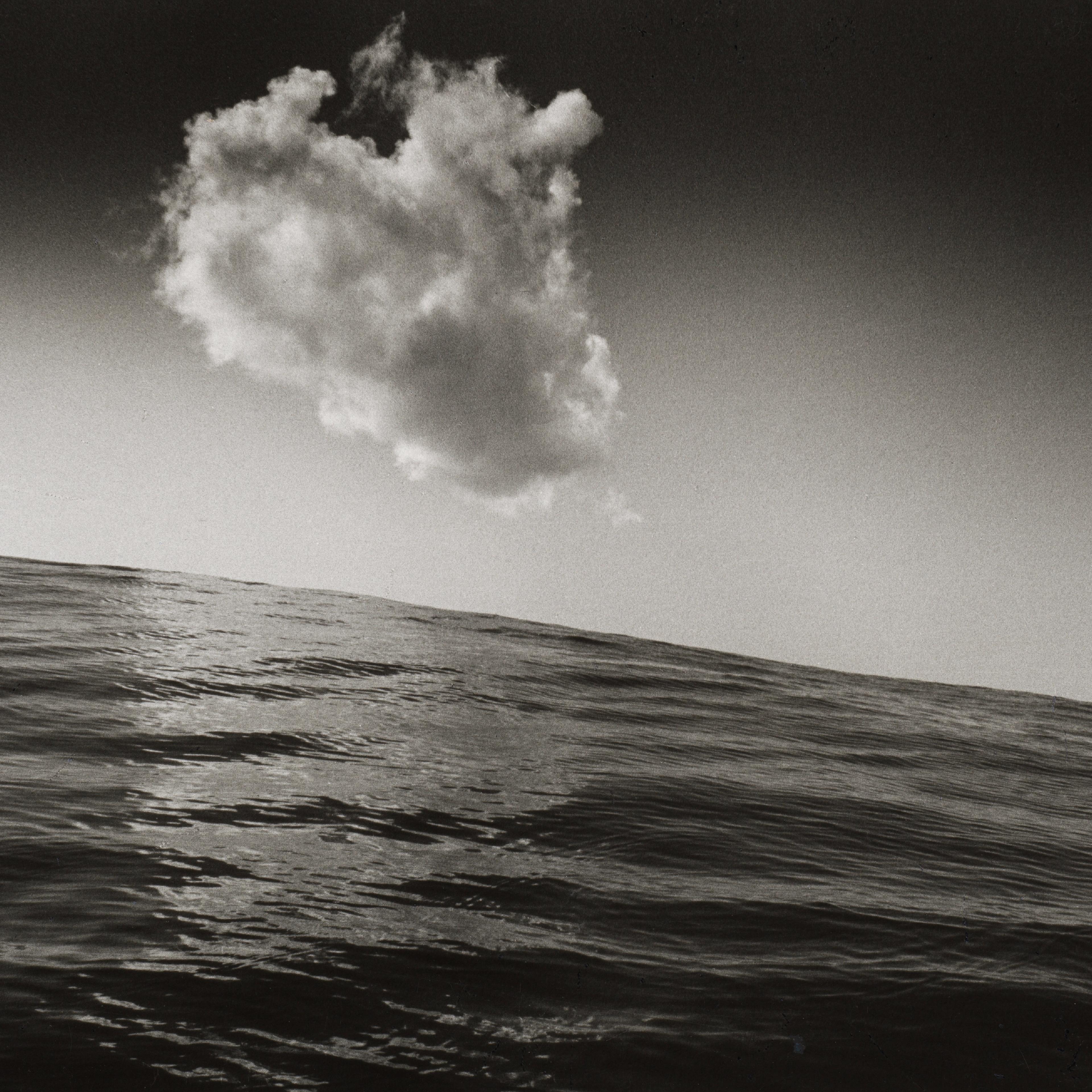 Black-and-white seascape with a large, billowing cloud above a slanted ocean horizon; textured waves and dramatic contrast .