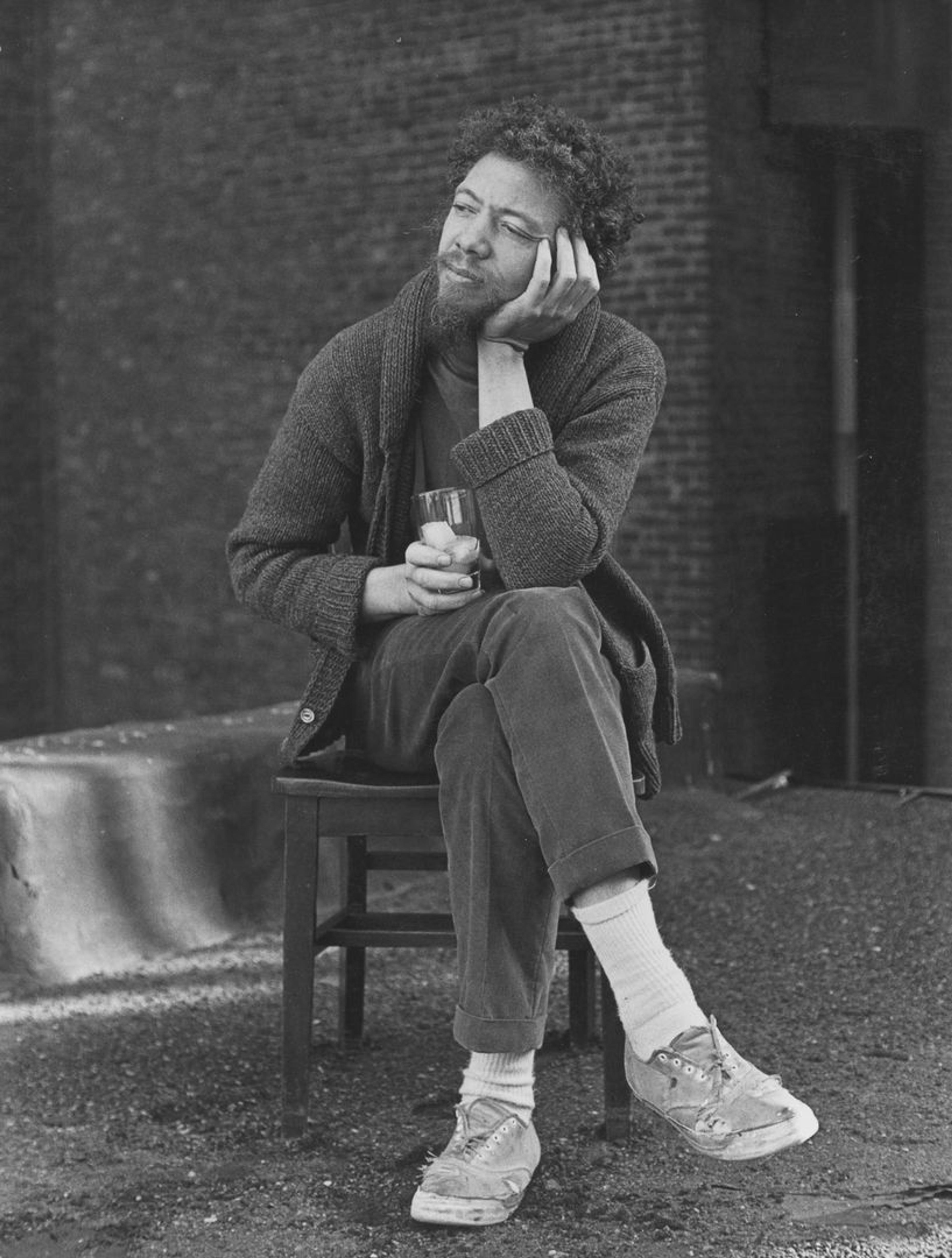 Black and white 1960s portrait of Benny Andrews with legs crossed, head resting in hands.