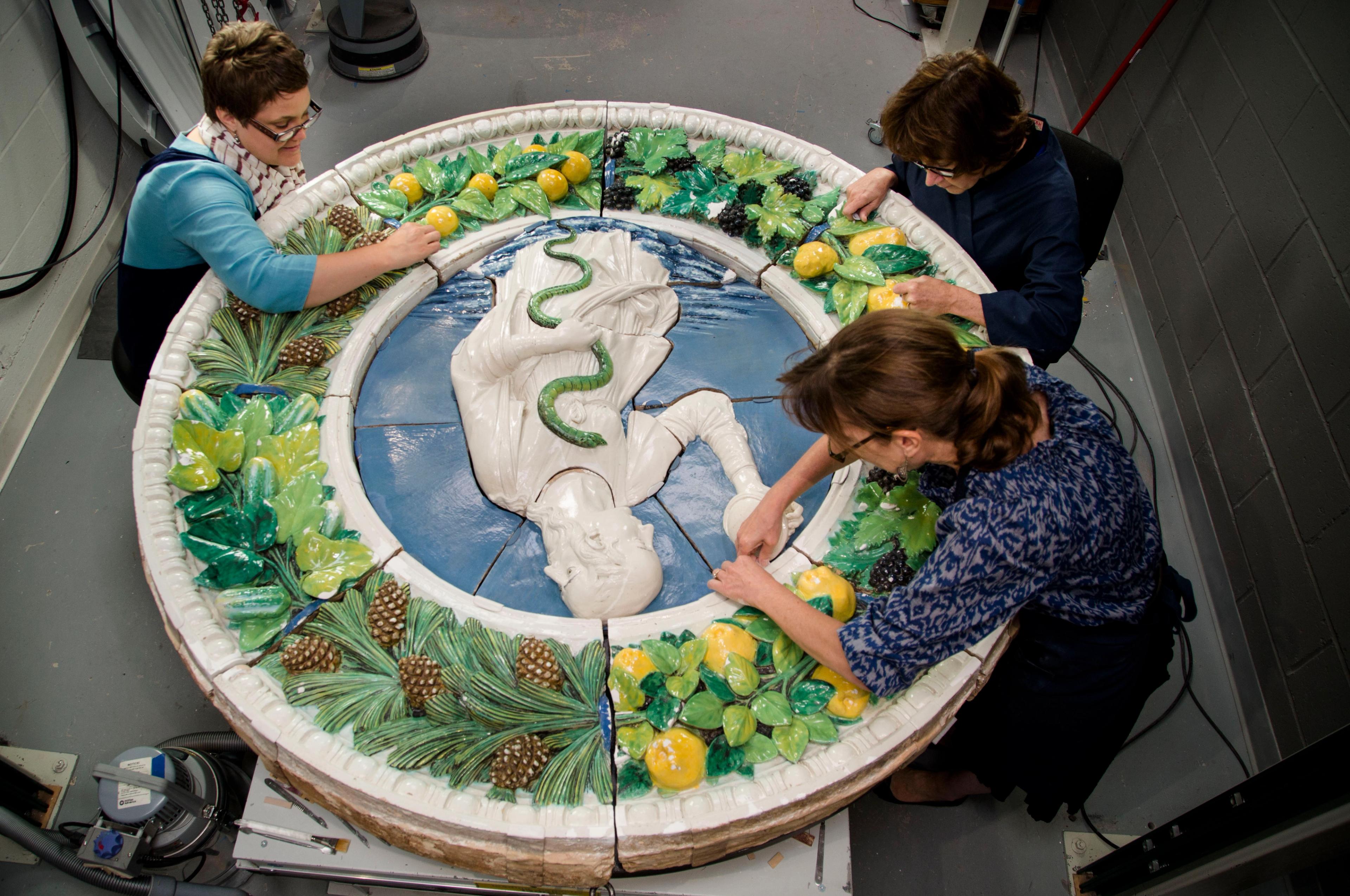 Three conservators working on a large, glazed, ceramic tondo with a human figure with a snake at center.