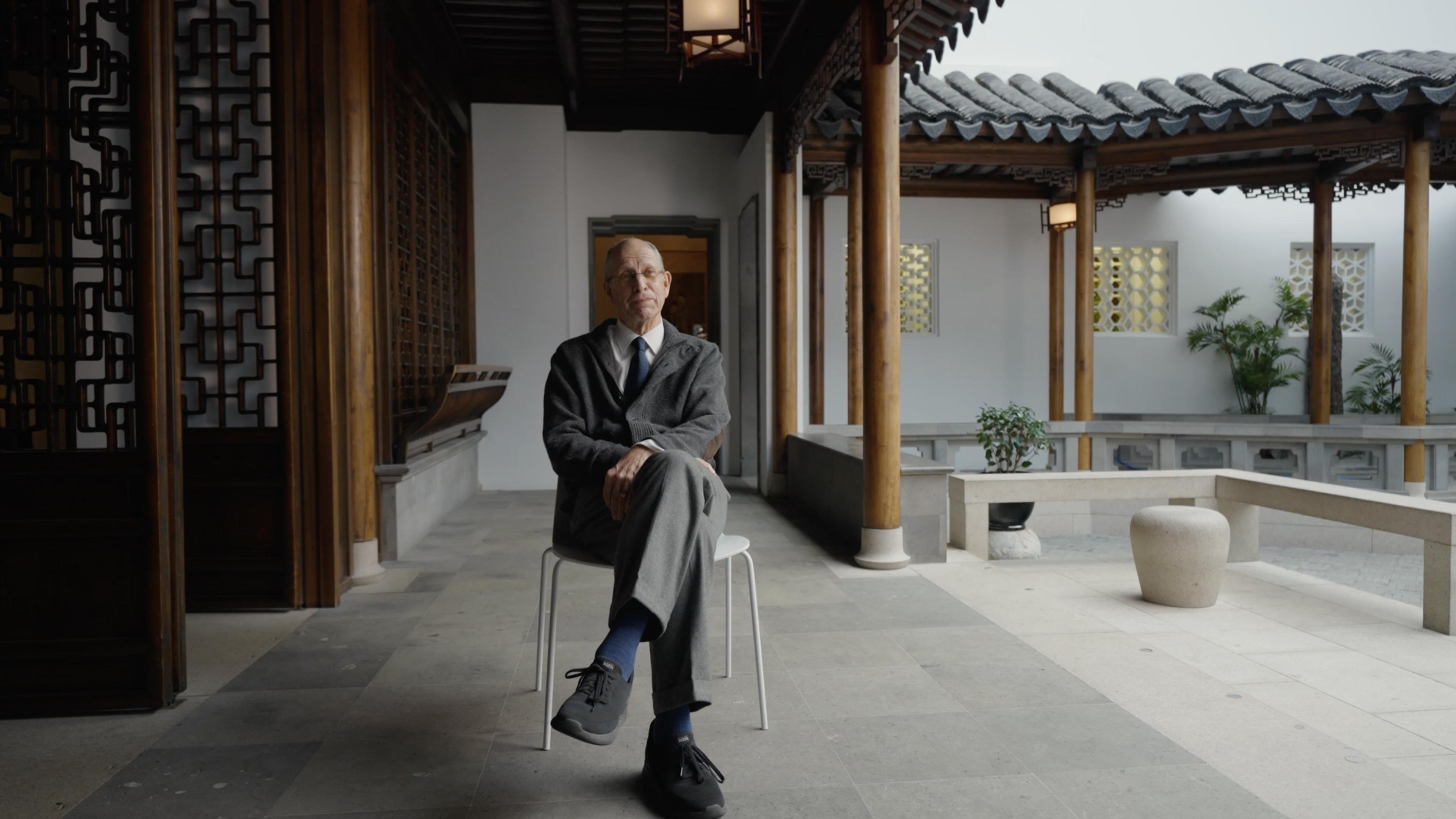 A man in office attire sits with his legs crossed in a brightly-lit, columned pavilion with geometric door insides and scalloped roof details.