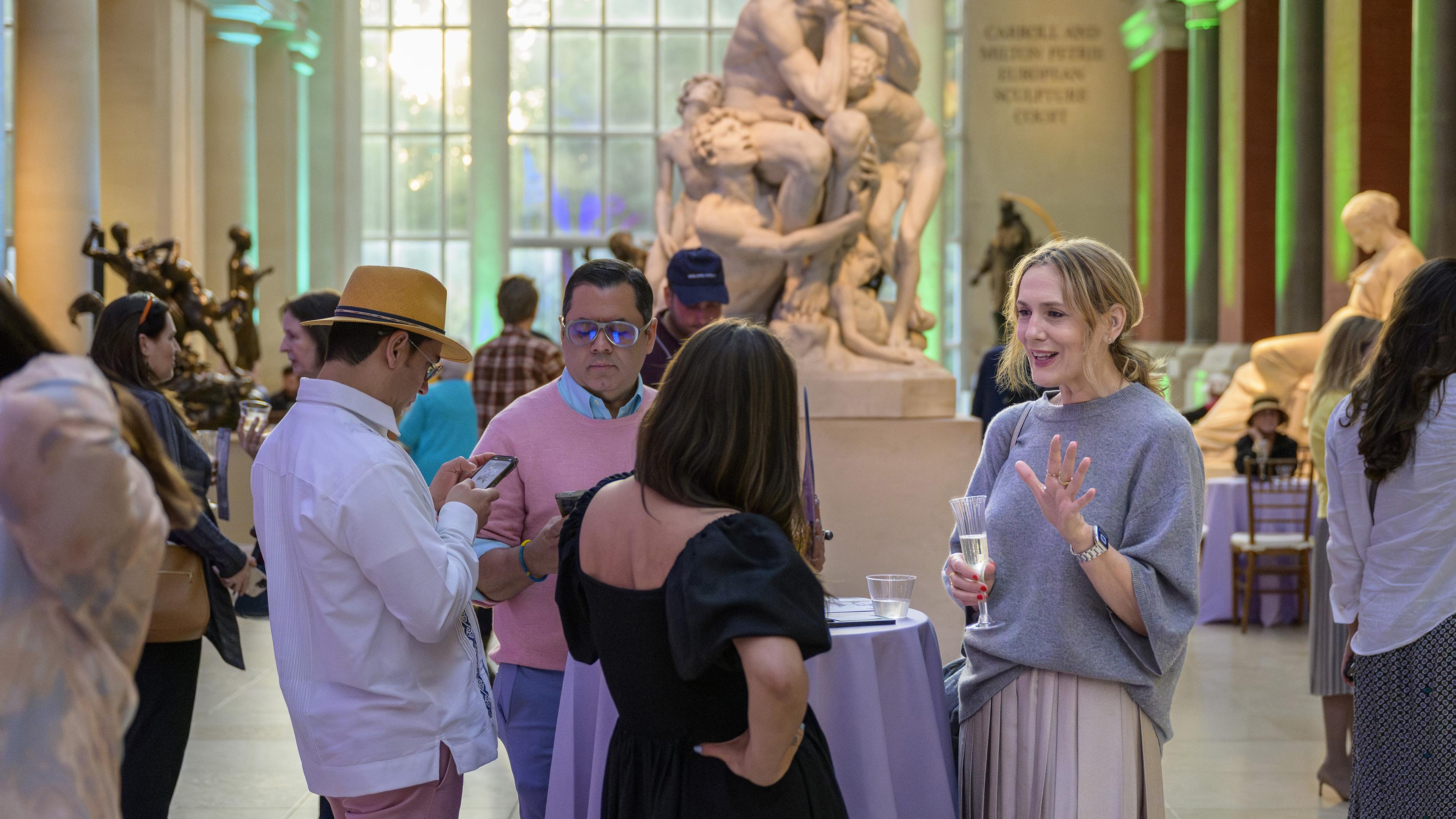 A group of four people stands around a table in The Met's Sculpture Court, talking and drinking cocktails.