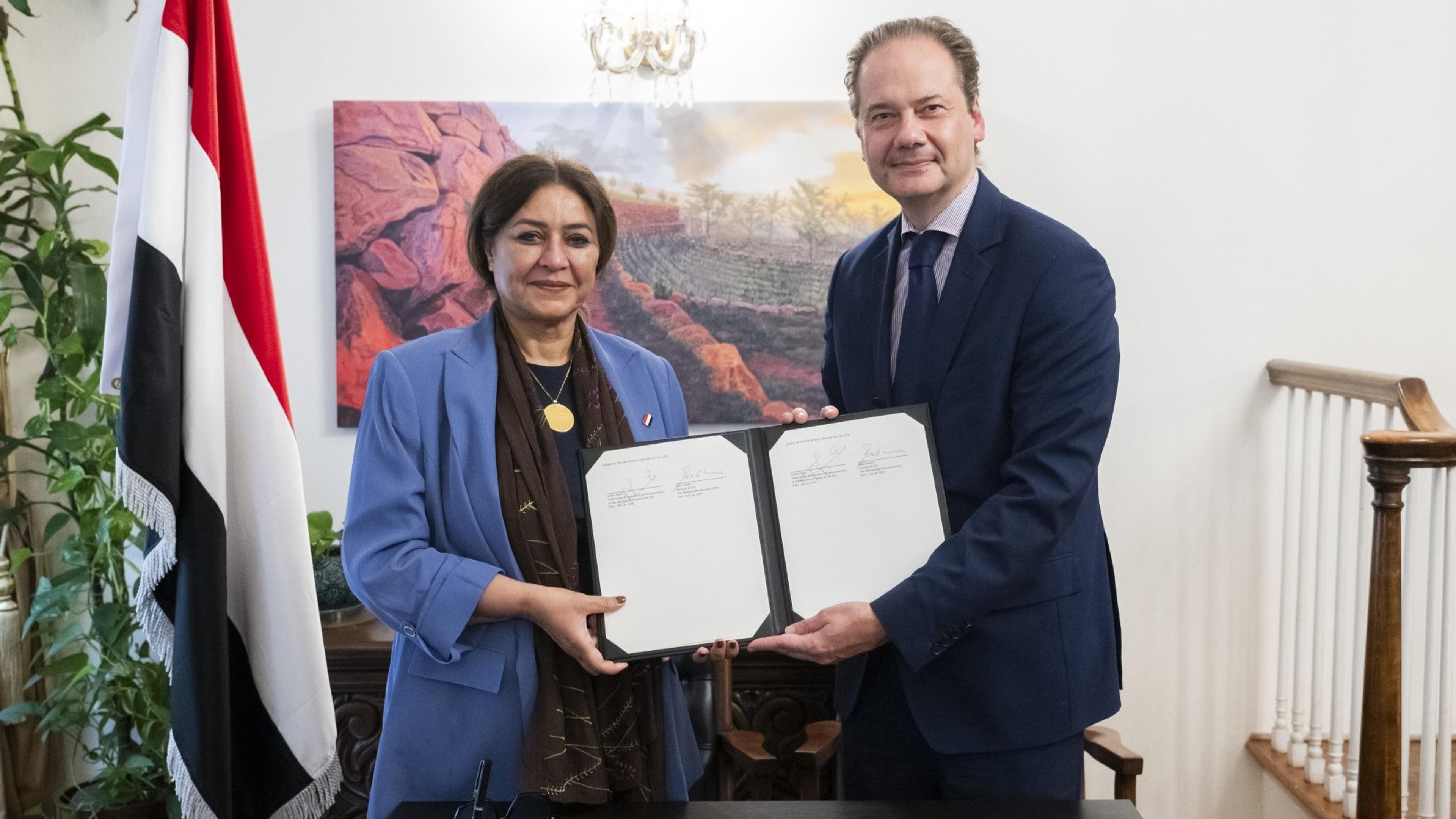 Two people holding a signed contract standing in front of a photograph of a sunny mountain