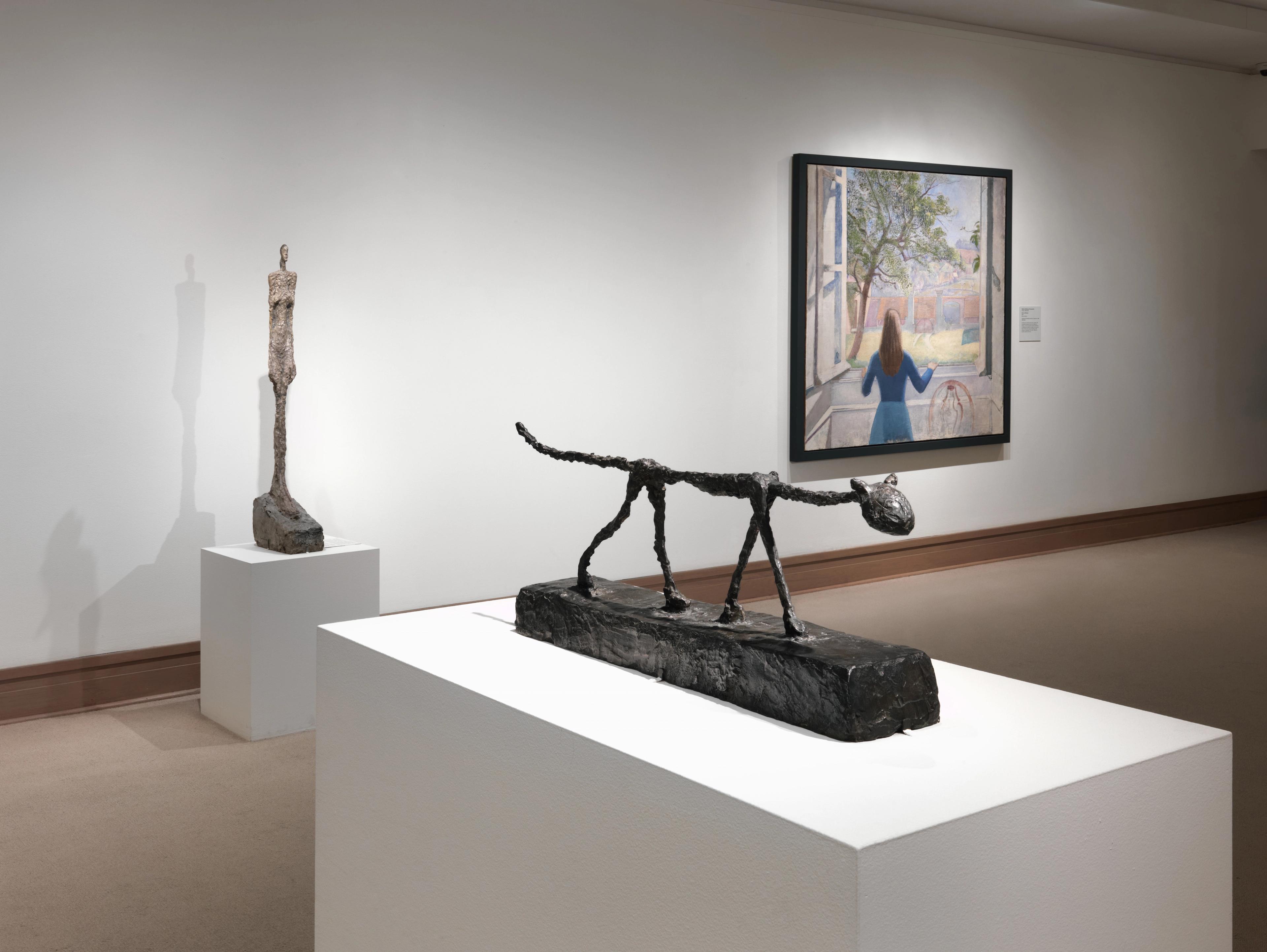Image of a museum gallery with a bronze sculpture of a stick-like cat on a pedestal in the foreground, a skinny bronze sculpture of standing a woman on another pedestal, and a painting on the back wall of a woman looking out a window with her back facing the viewer.