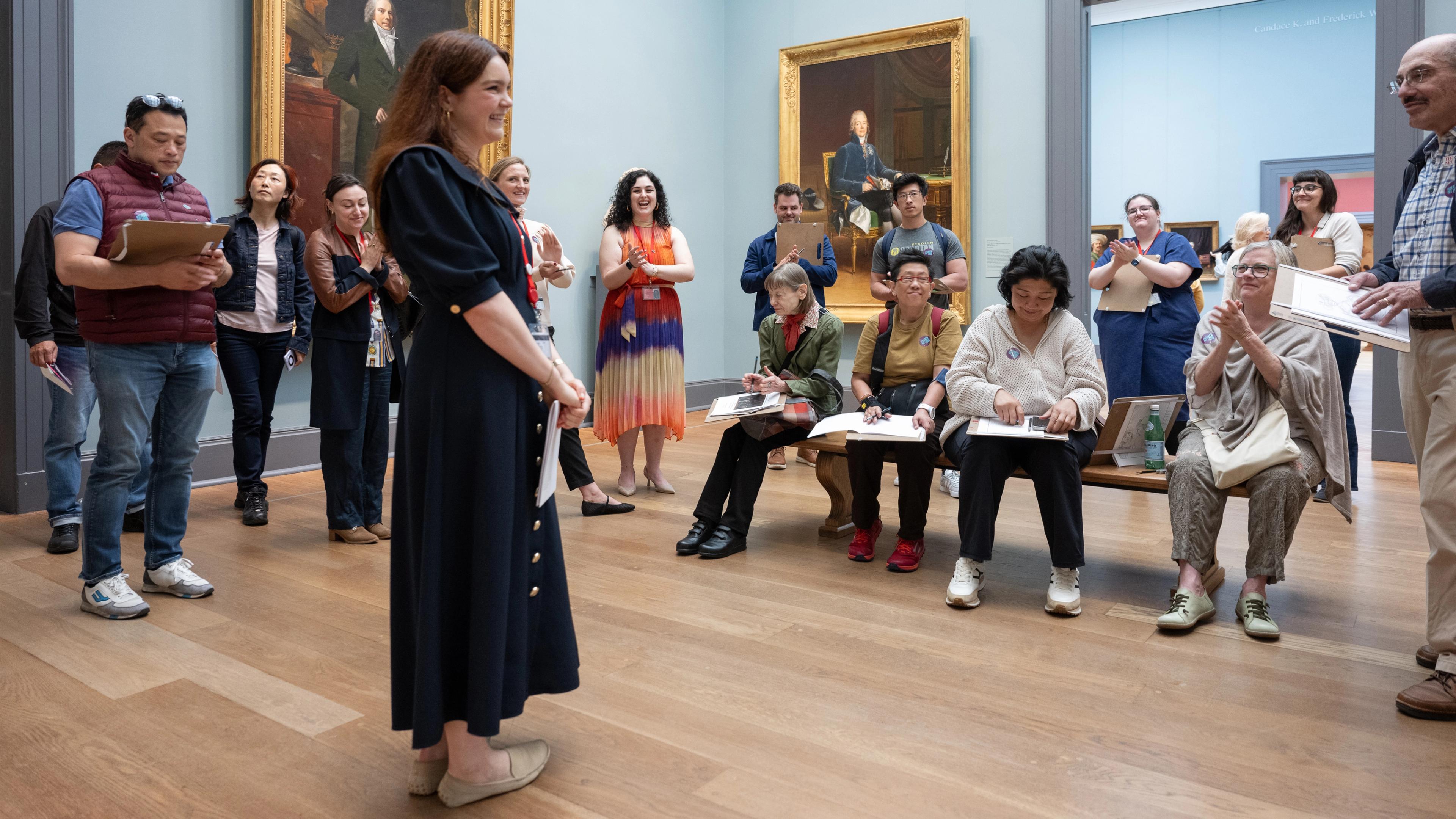 A woman in a long black dress is speaking to a group of people who are sketching in a museum gallery with large classical paintings on the walls.
