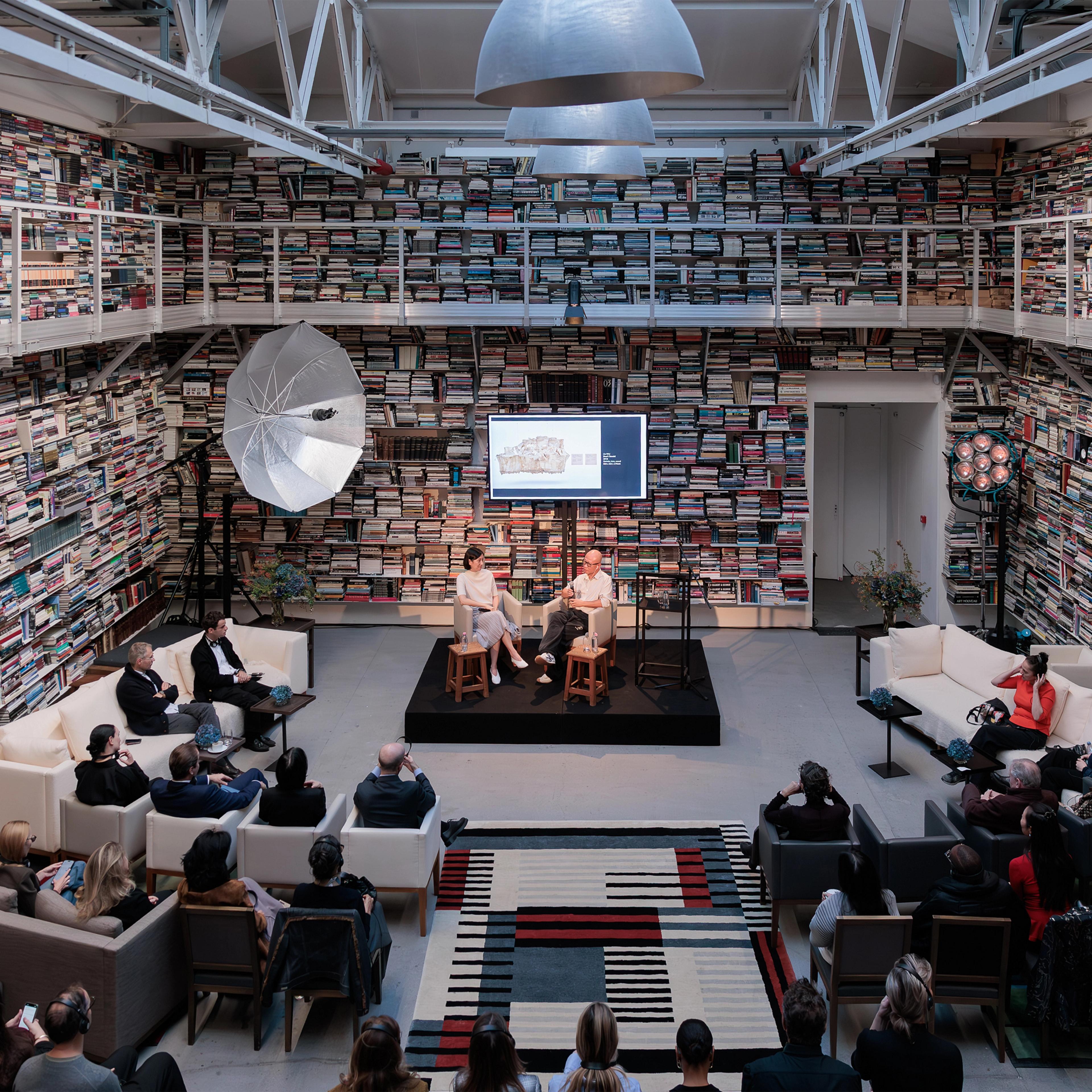Two speakers seated on a small raised platform surrounded by books, while an audience listens attentively in a cozy setting.