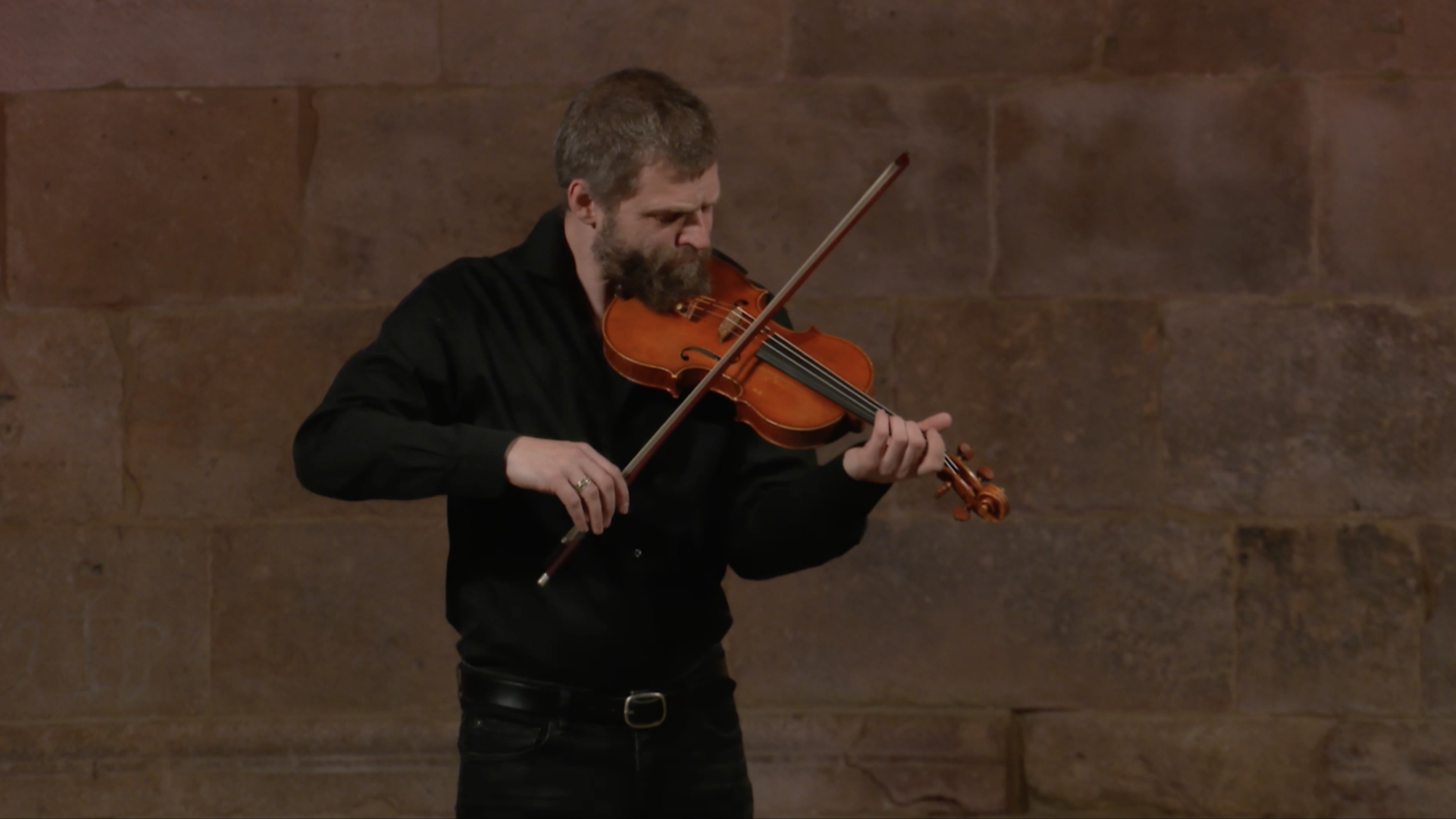 Man on stage, standing tall, wearing all black as he plays his violins