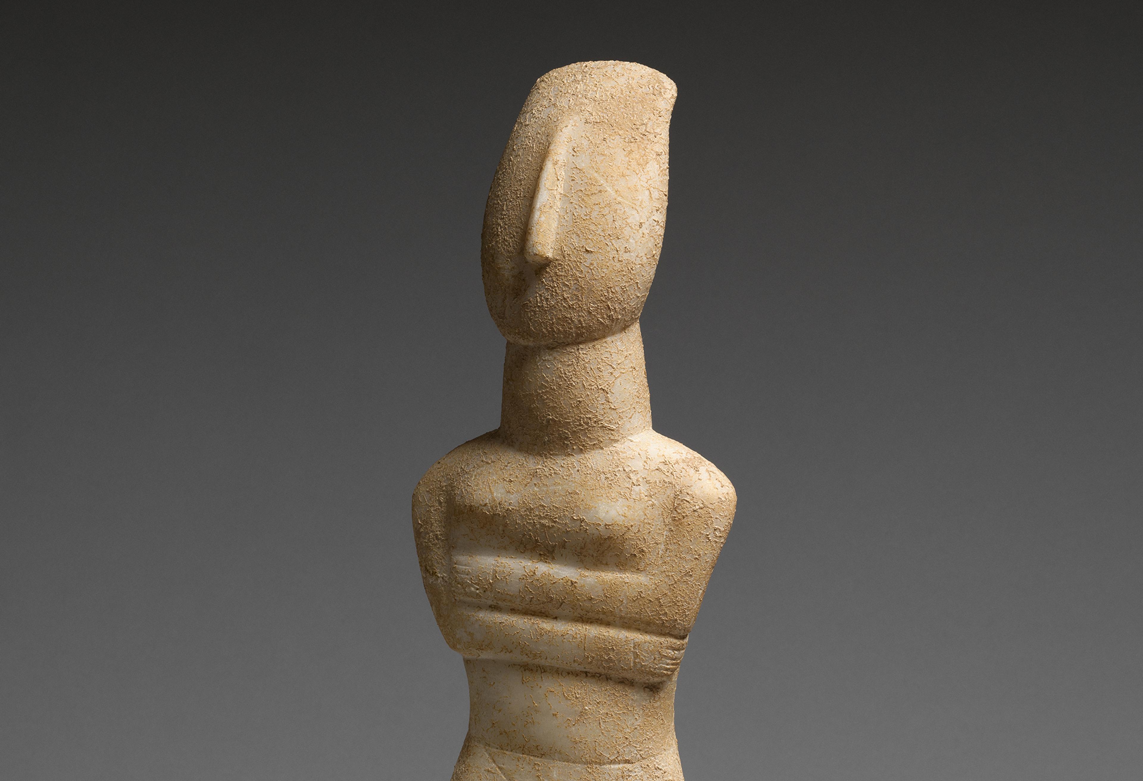 Nude stone Cycladic figure with arms crossed against a grey background.