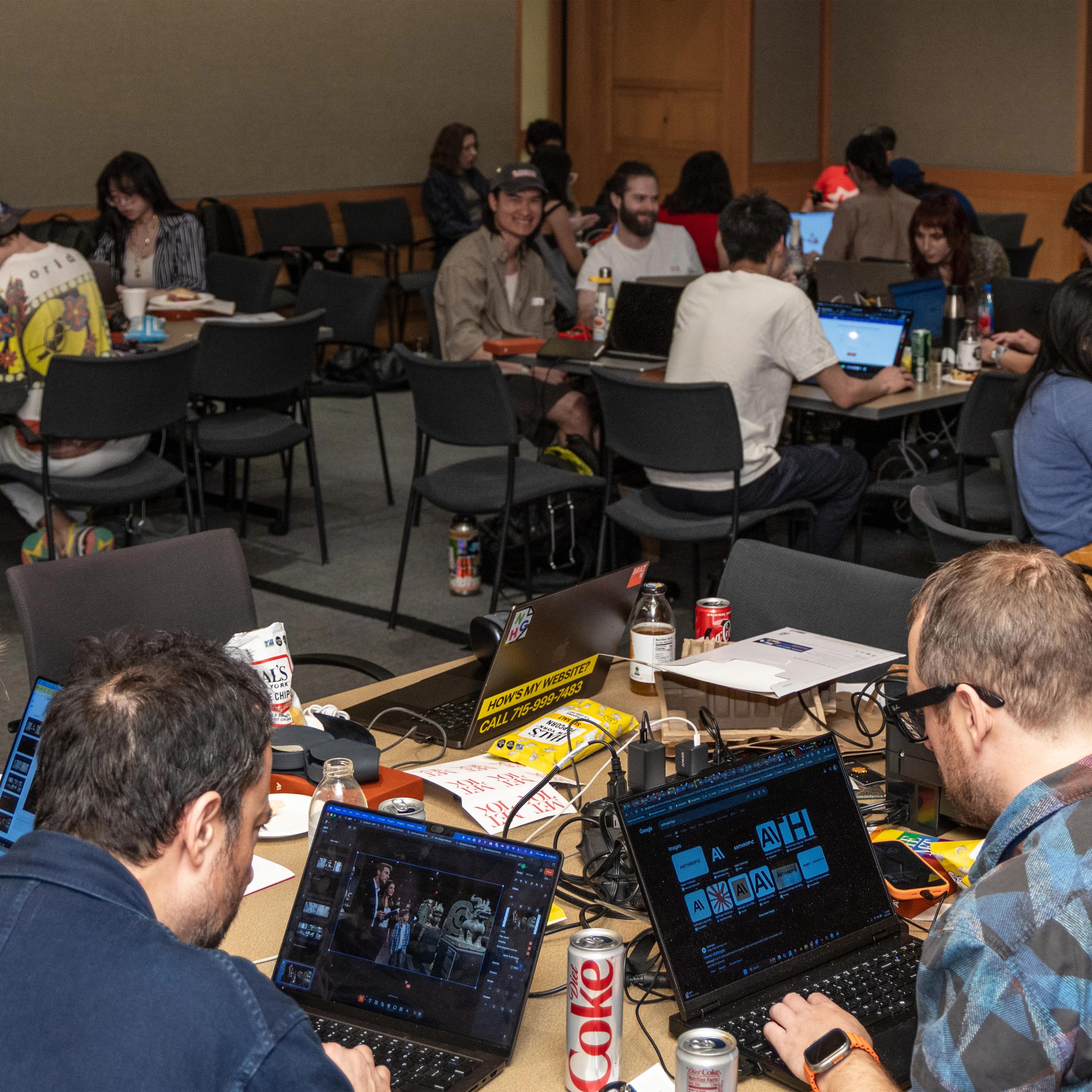 A group of people in a conference room are engaged in collaborative work on laptops. The atmosphere is focused and lively, with drinks and snacks on tables.