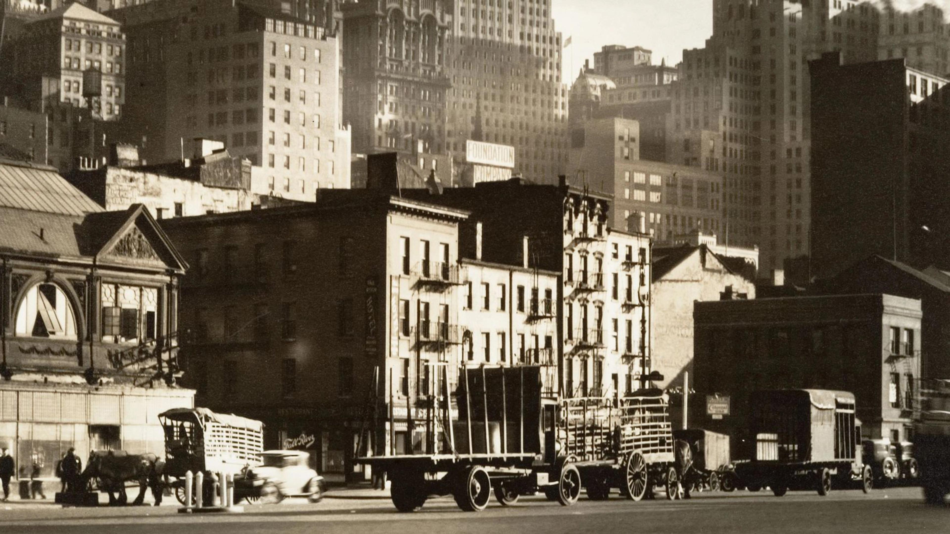 Sepia-tone photograph of old-timey wagon trucks and cars traveling up and down West Street in New York City set against a backdrop of multi-story apartments and midsize skyscrapers.