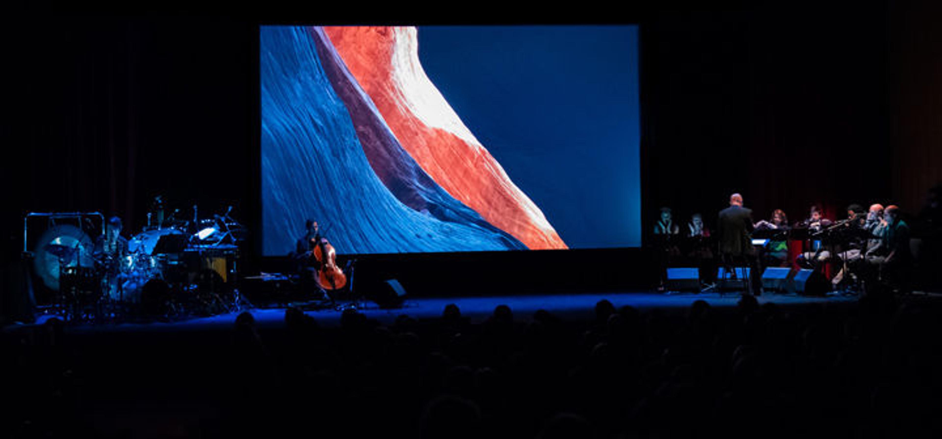 On a darkened stage several musicians and a chamber choir perform before a screen projection of a film depicting the Colorado River basin